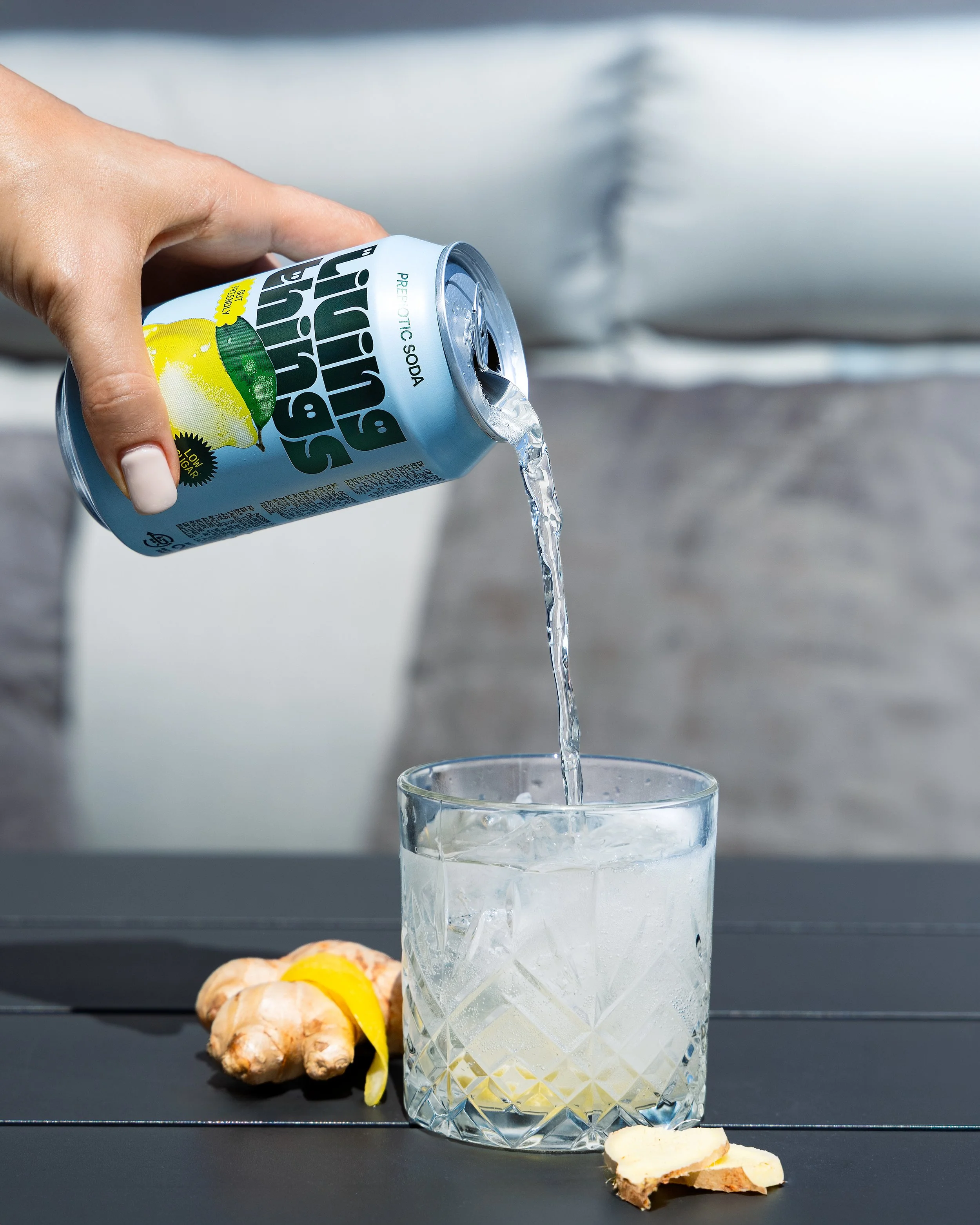 A hand pouring lemon soda from a can into a glass with ice. Ginger root and lemon slices are on a dark surface in the background.