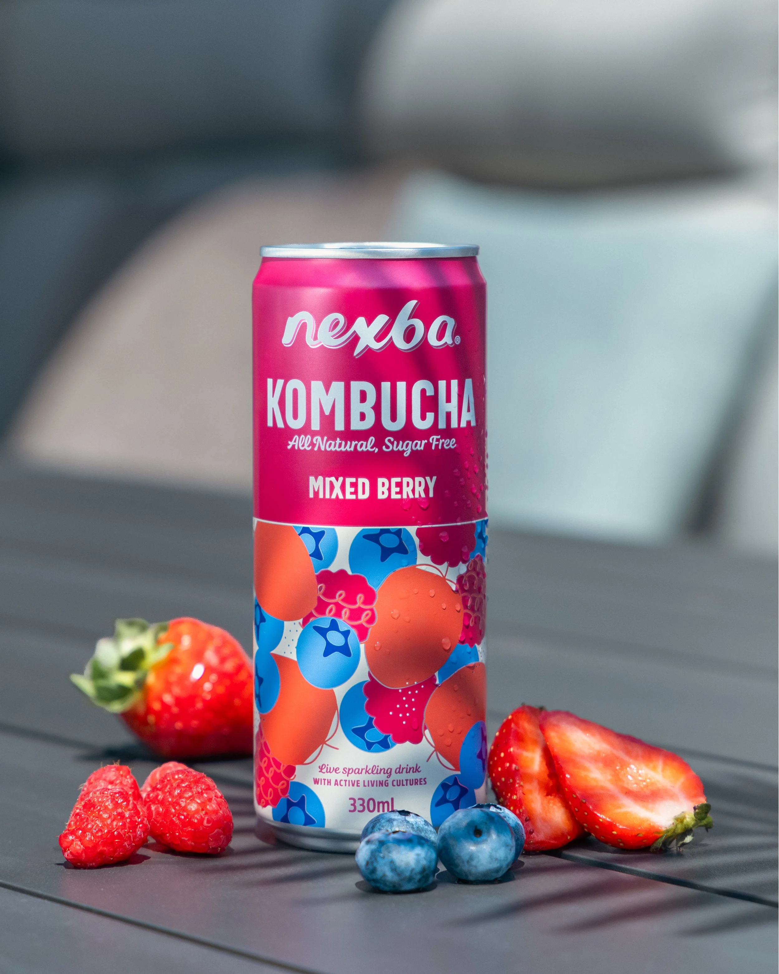 A can of Nexus Kombucha flavored with mixed berries is placed on a dark surface, with fresh strawberries, blueberries, raspberries, and sliced strawberries surrounding it. The can is pink and decorated with images of berries, with text indicating it is a live sparkling drink with active living cultures, 330ml.
