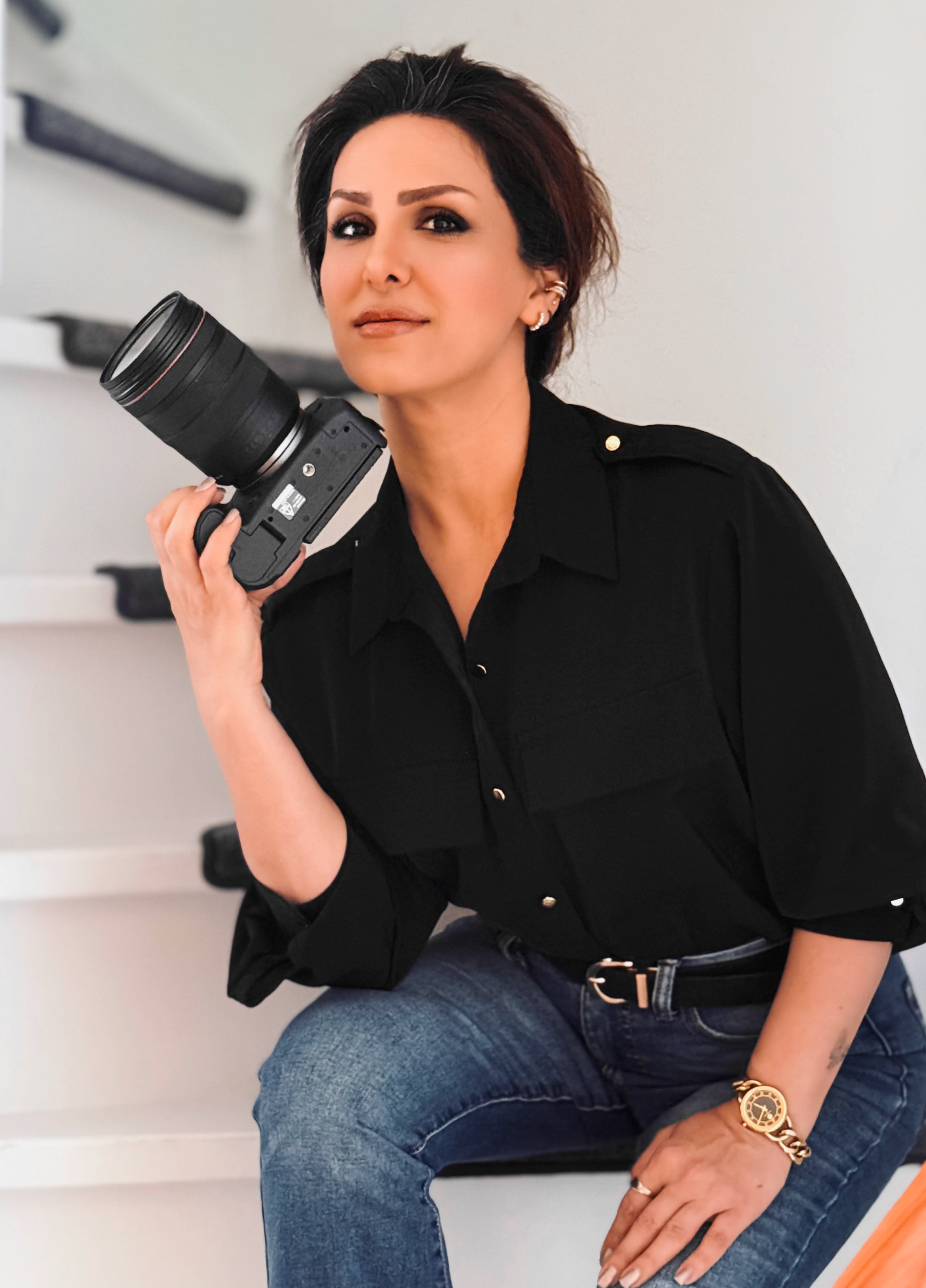A woman with short dark hair, wearing a black shirt and blue jeans, holding a camera with a large telephoto lens, sitting with one knee up and her hand resting on it, in a room with white walls and framed photographs.