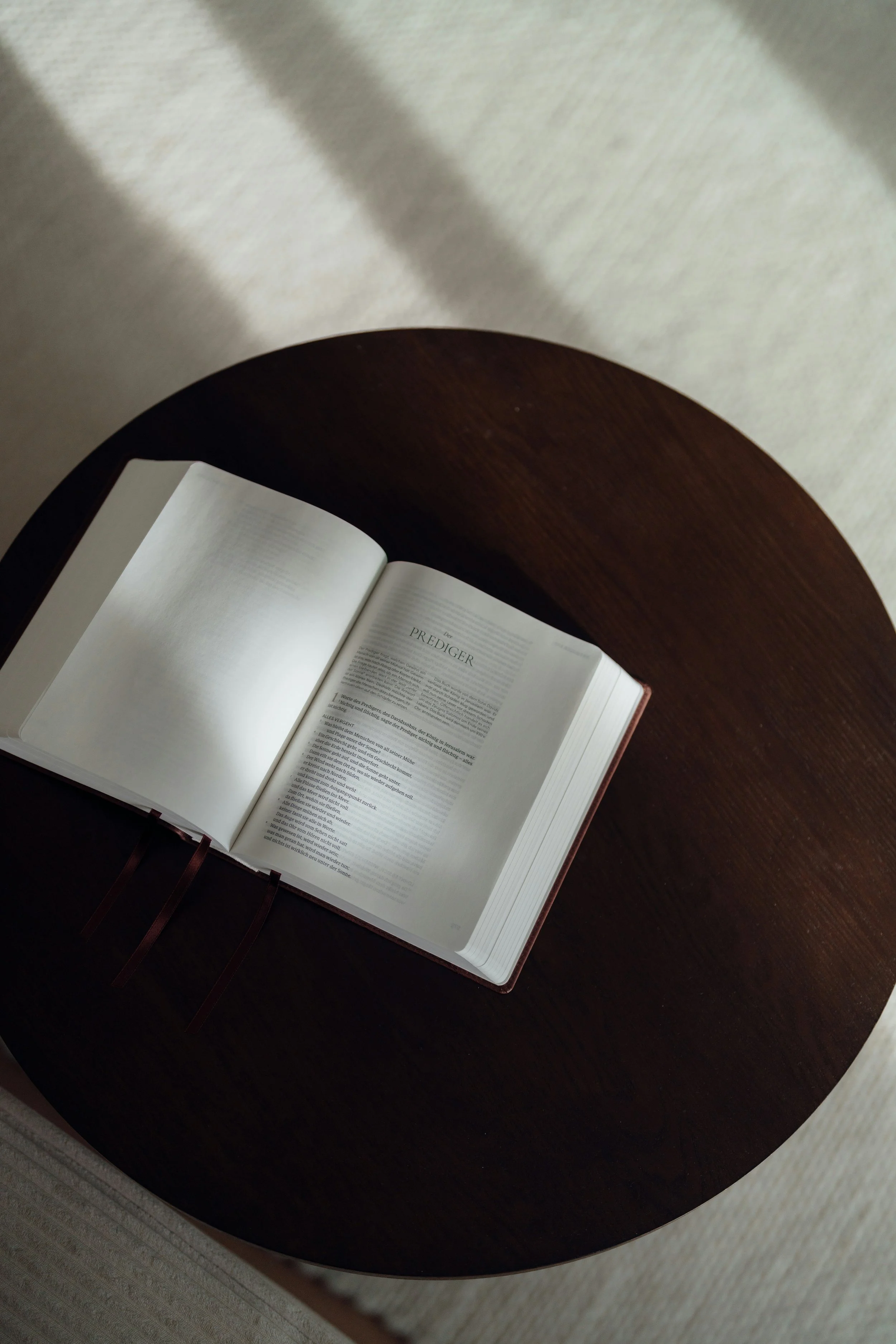 An open book on a dark wooden table, with a page titled "Prediger" visible, lit by natural light creating shadows on the surface.
