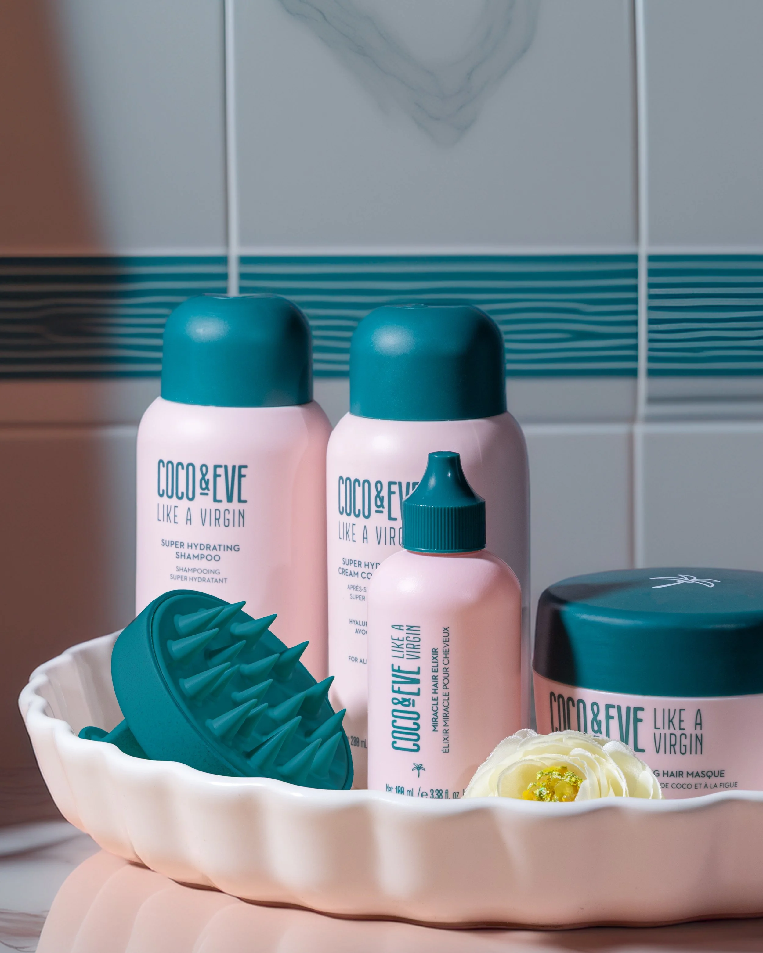 Set of Coco & Eve hair care products including a super hydrating shampoo and a cream coconut conditioner, placed in a decorative bowl with a teal scalp massager and a flower.