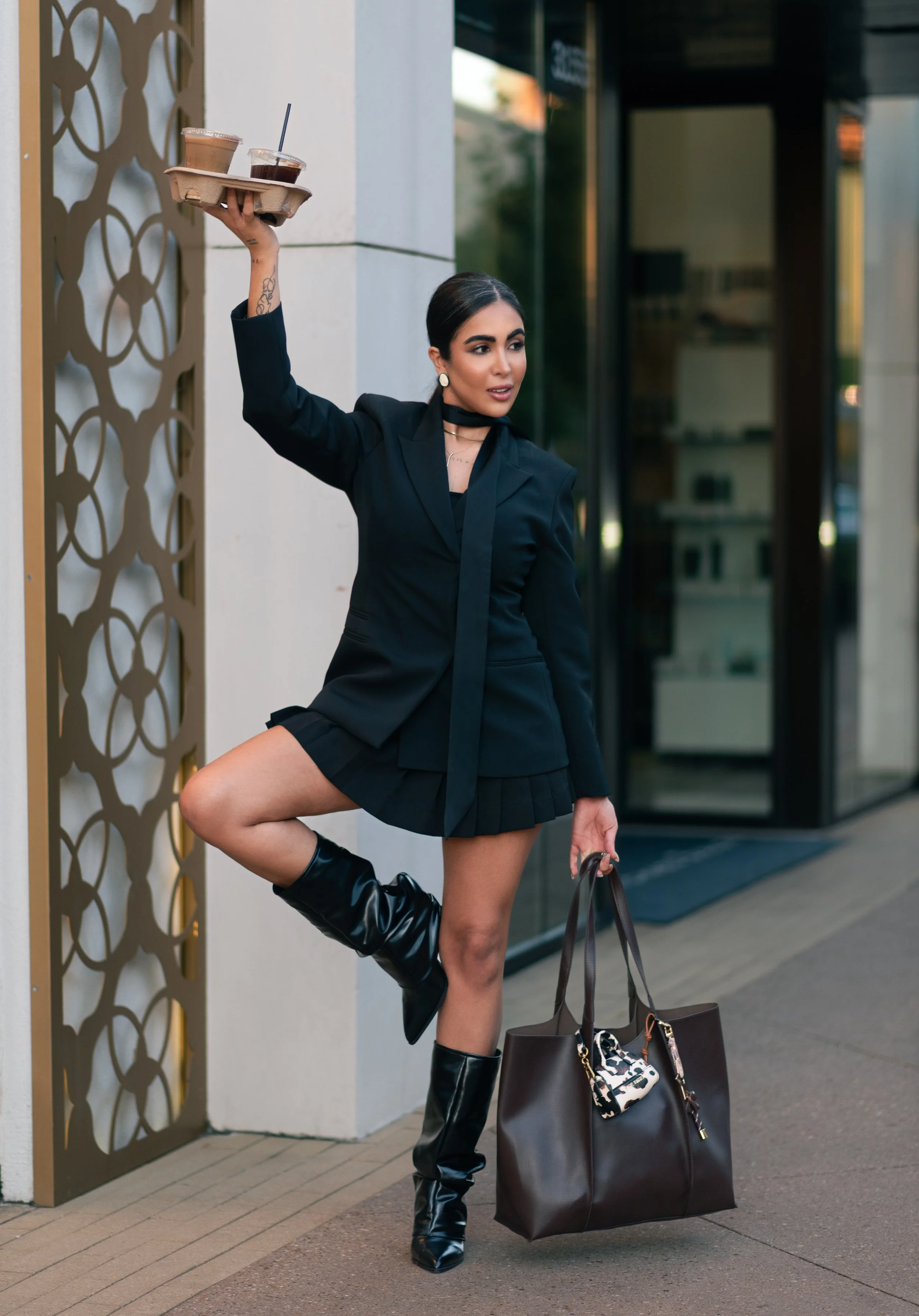 A woman in a black blazer and pleated skirt outside a building, holding a tray with two drinks, wearing black knee-high boots, and carrying large brown and patterned handbags.
