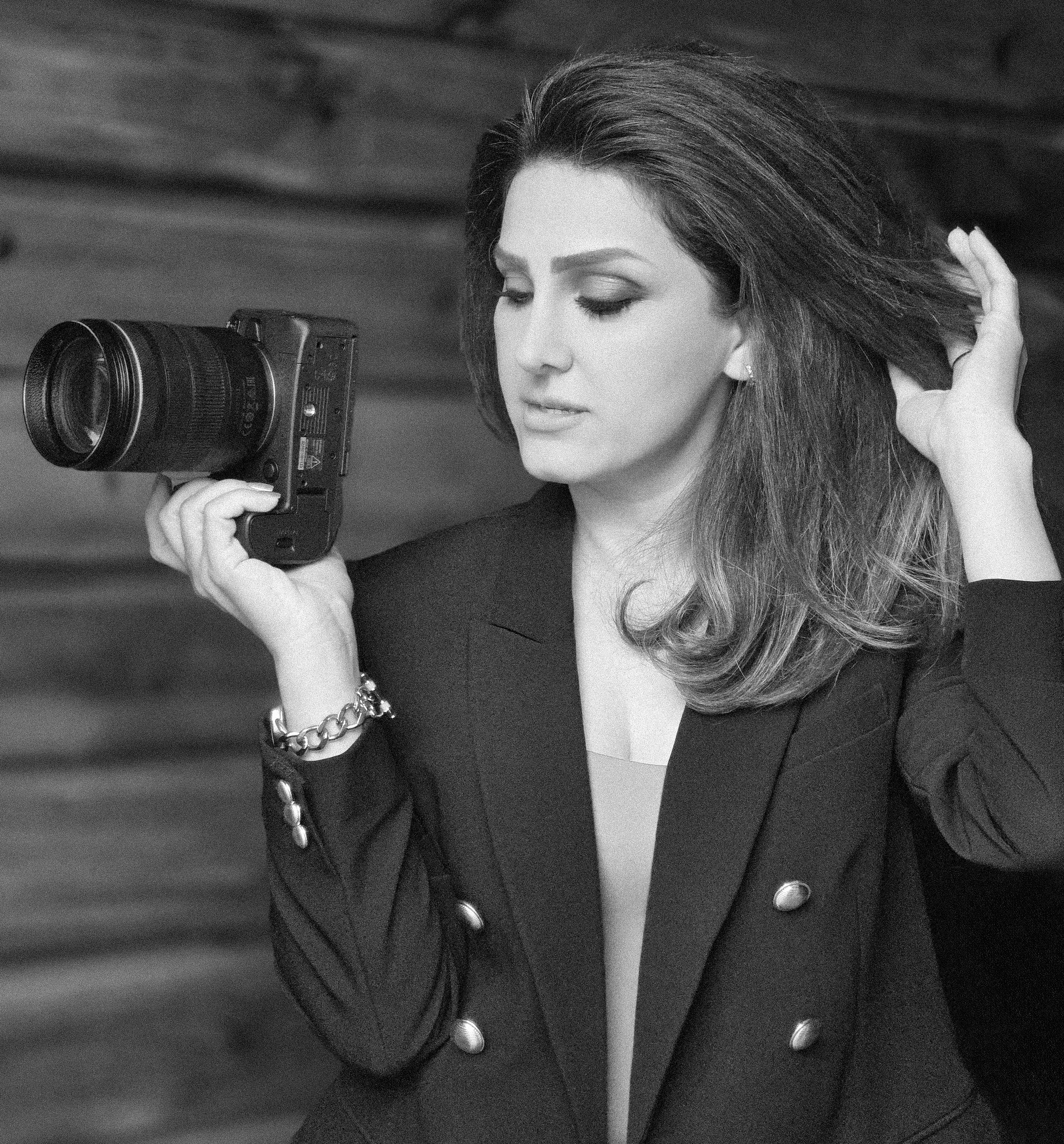 Black and white photo of a woman with long hair, wearing a dark blazer with pearl buttons, jewelry, and a bracelet, holding a camera and touching her hair.