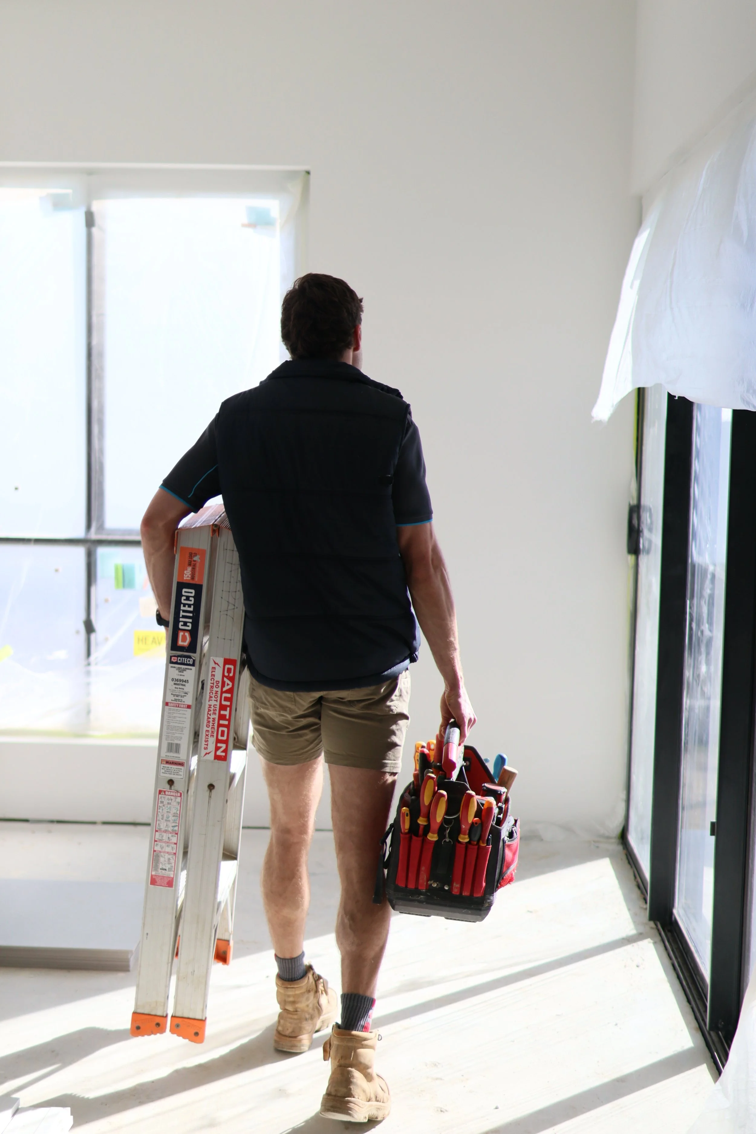 A man walking inside a house construction site, carrying a toolbox and a ladder, with sunlight streaming through the windows.