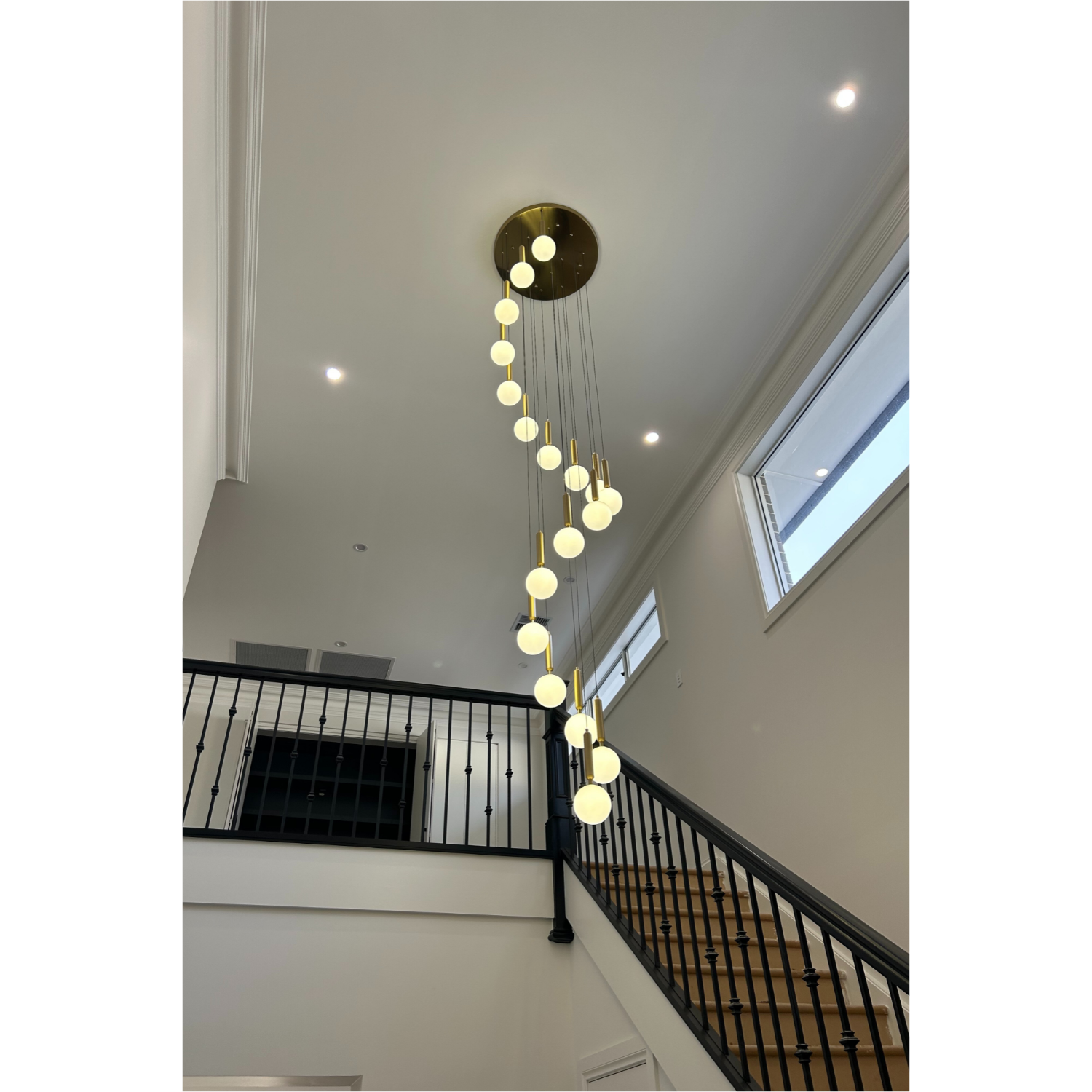 Modern chandelier with multiple evenly spaced spherical lights hanging from a gold fixture in a high-ceiling foyer with staircase and large windows.