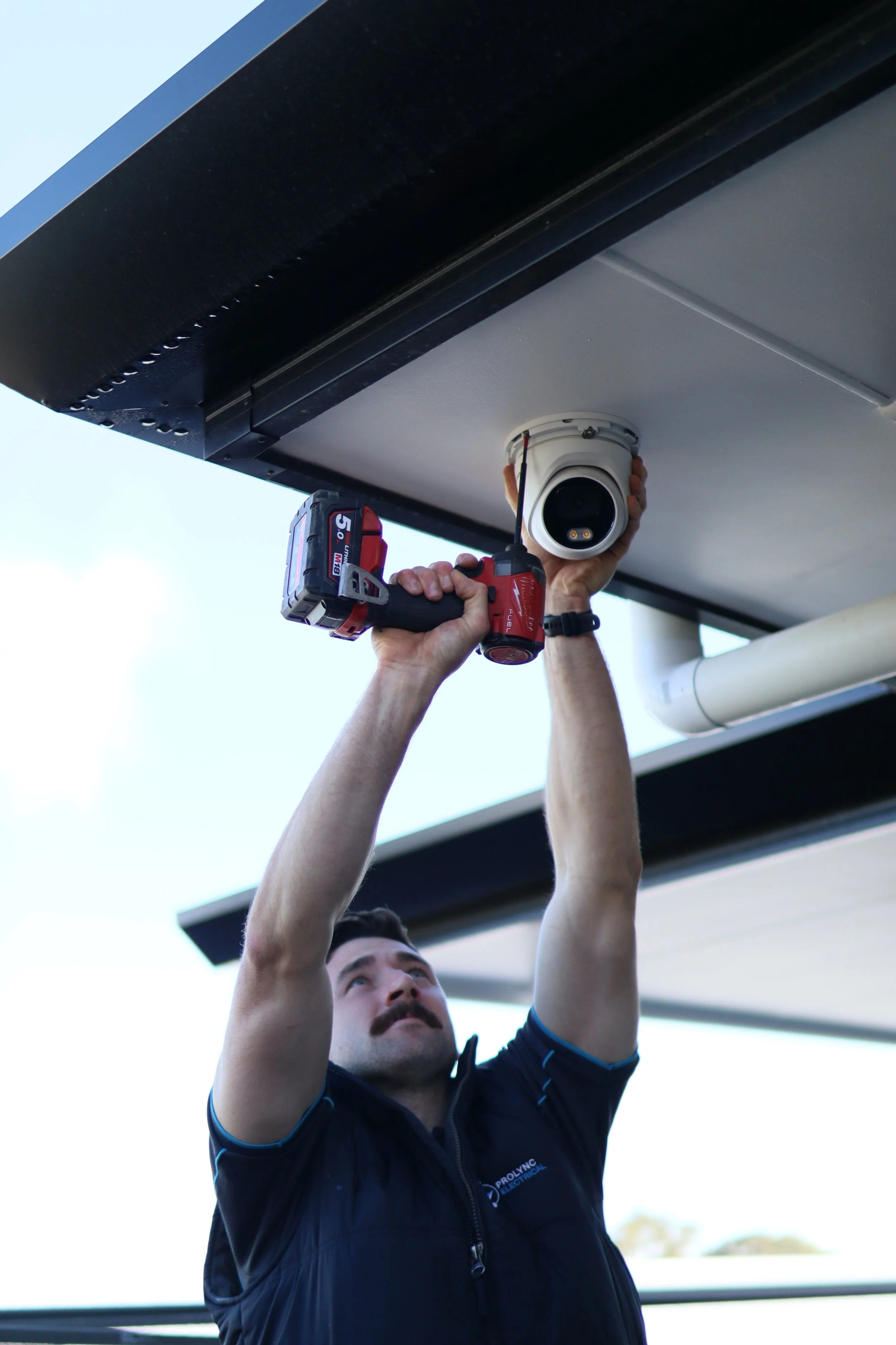 Man installing or repairing a security camera under an overhang with a cordless drill.