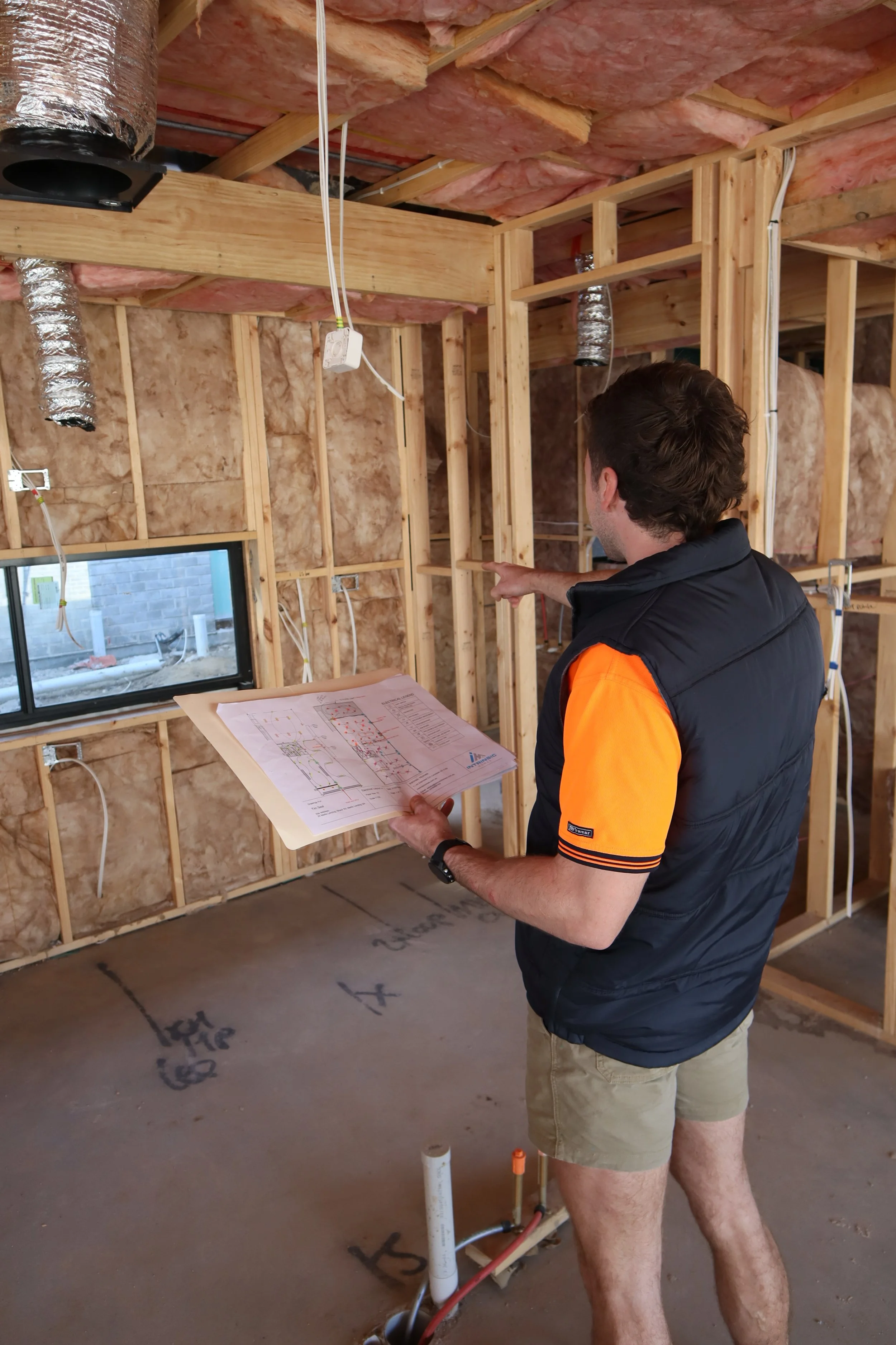 A man inspecting a construction site inside a house under renovation, holding a blueprint and pointing at the wooden framing of the wall.