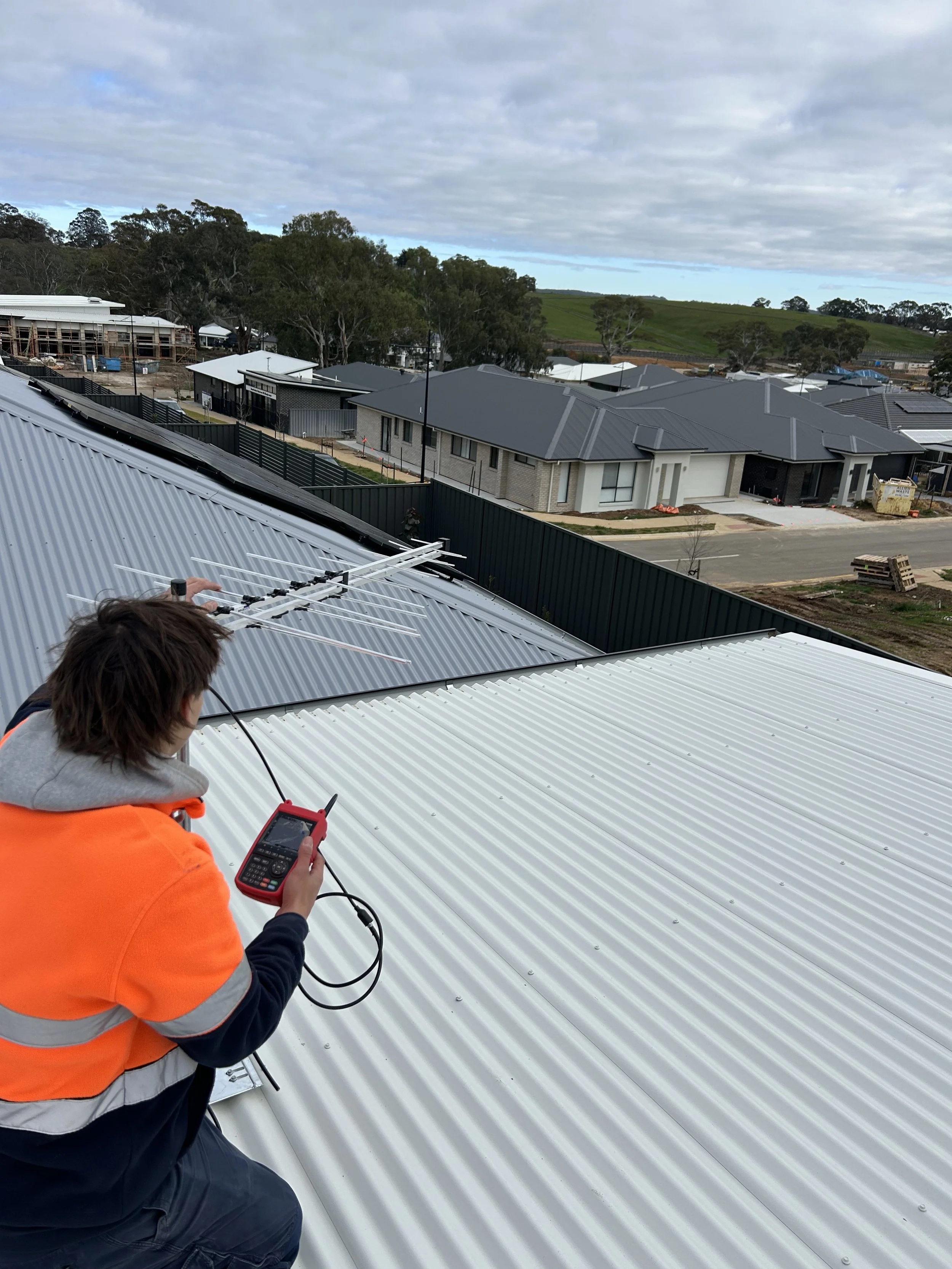 A worker in an orange safety vest and dark clothing is on a metal roof, using a handheld device to conduct test on an antenna. The background shows a residential area under construction with houses, trees, and a cloudy sky.