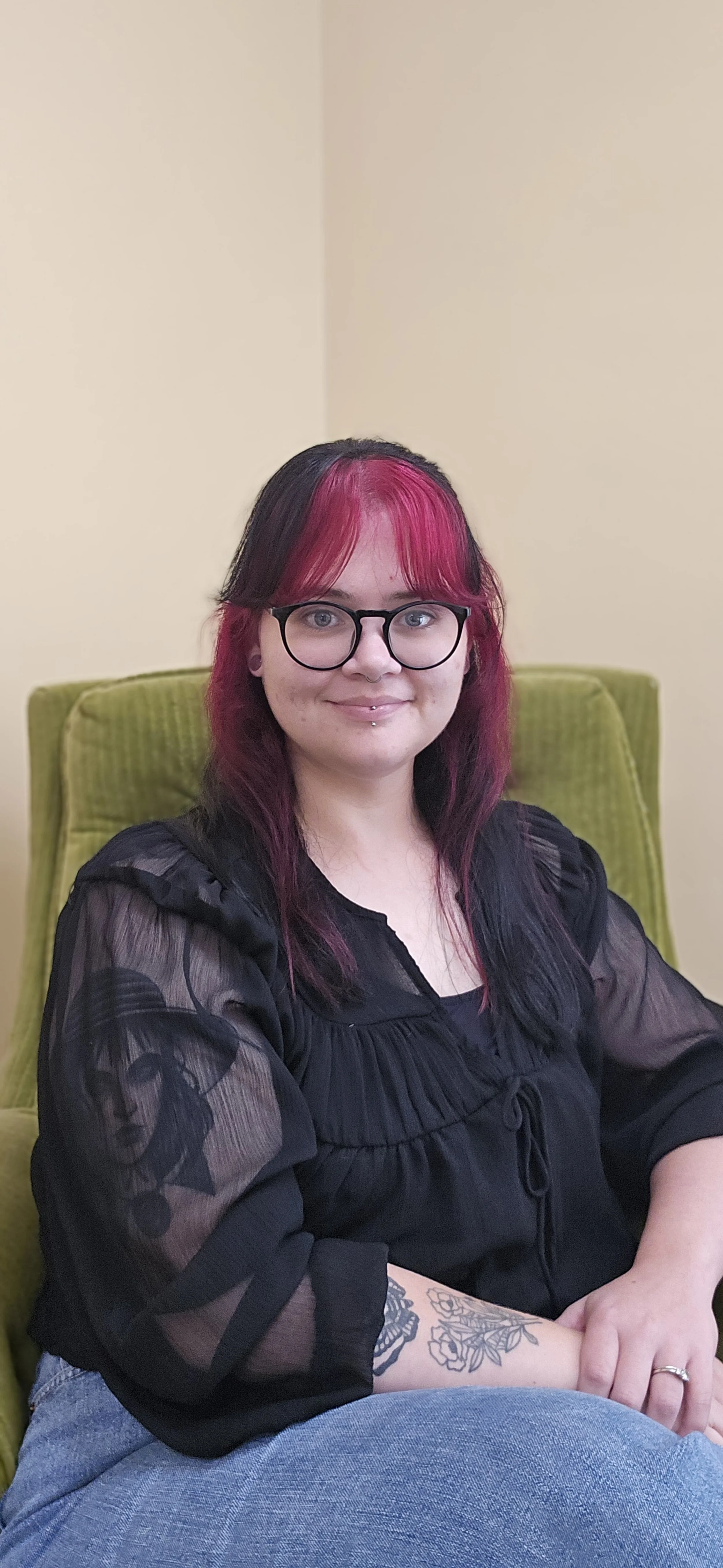 A woman with pink and black hair, wearing glasses, sitting on a green chair with a light-colored wall in the background.