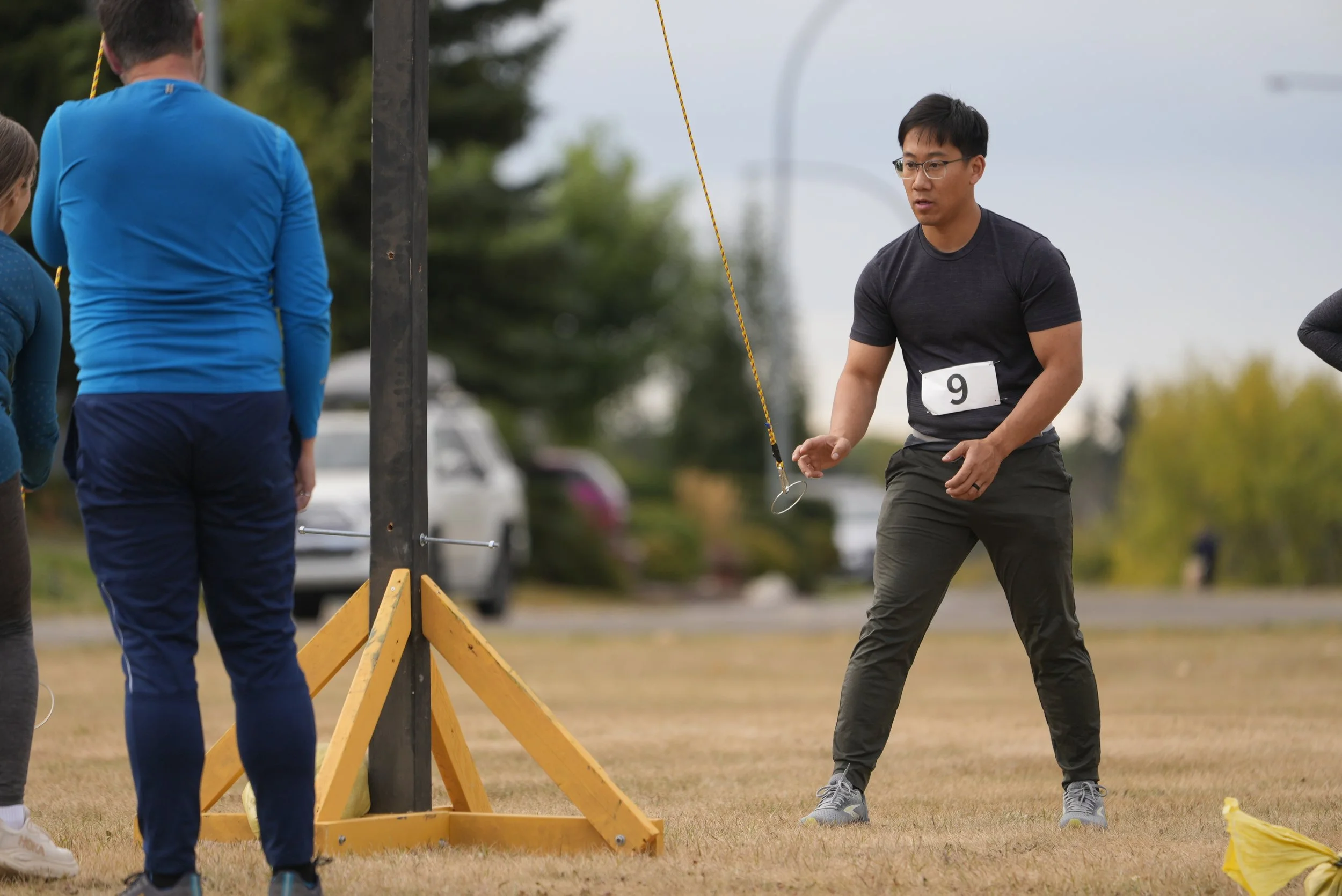 A man wearing glasses and a black t-shirt with the number 9 on it is participating in an outdoor activity that involves throwing a ring at a target. Other people are visible nearby, and the scene is set on a grassy field with trees and parked cars in
