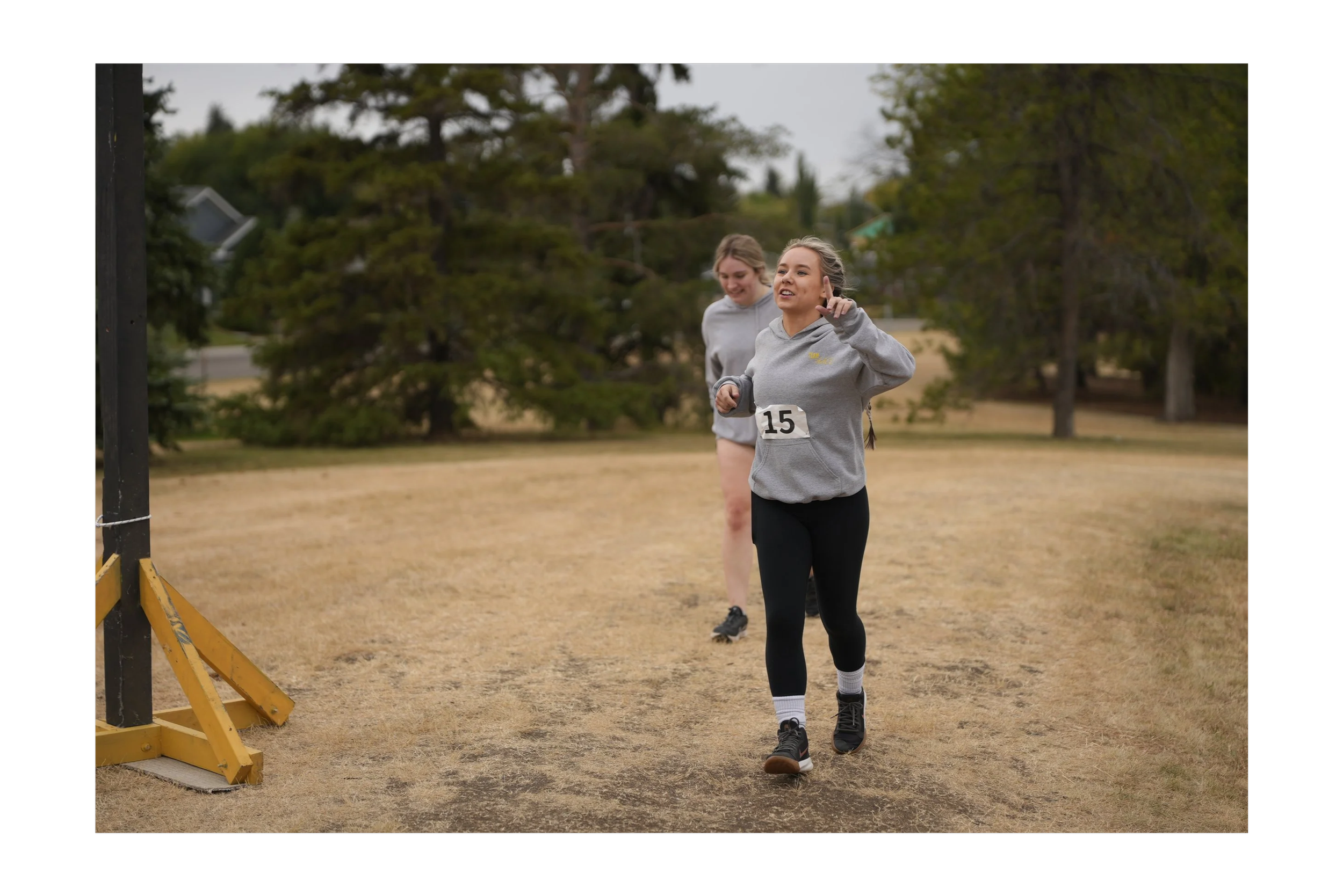 Two women running outdoors near a fenced area with trees in the background. One woman, dressed in gray with a number 15 on her shirt, is smiling and pointing, while the other woman is slightly behind, also in athletic wear.