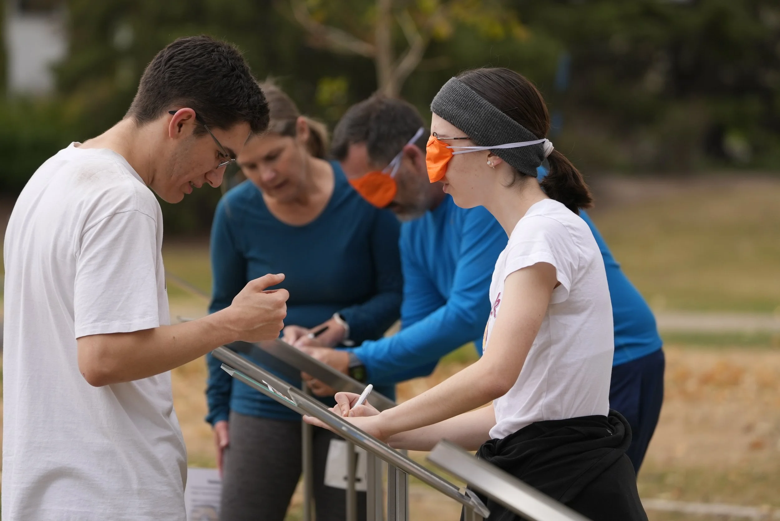 A group of people outdoors, with a woman wearing a blindfold and orange nose cover, engaged in an activity with a guide. The woman and guide are facing each other, with other participants in the background observing.