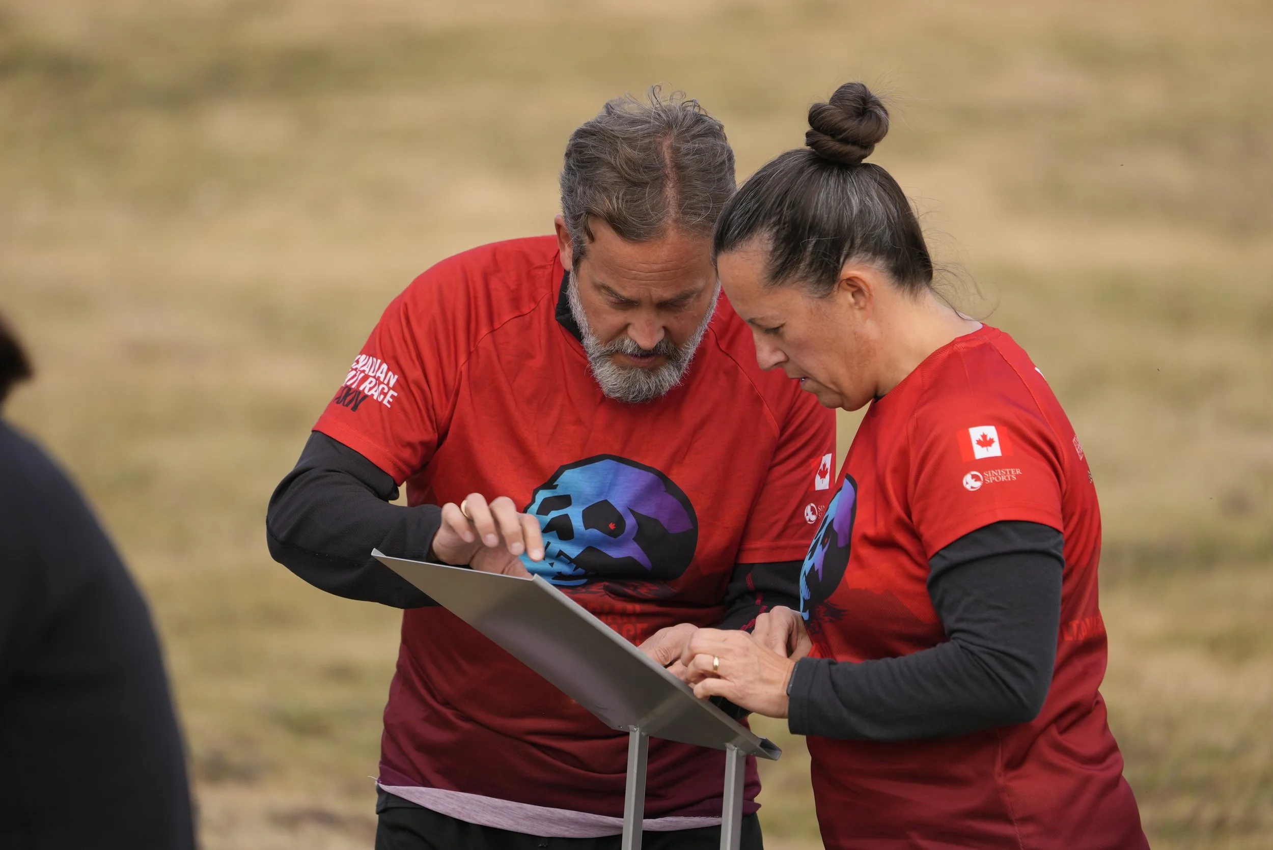 Two women and a man, all wearing red shirts with a Canadian flag patch, look at a tablet outdoors on a grassy field.
