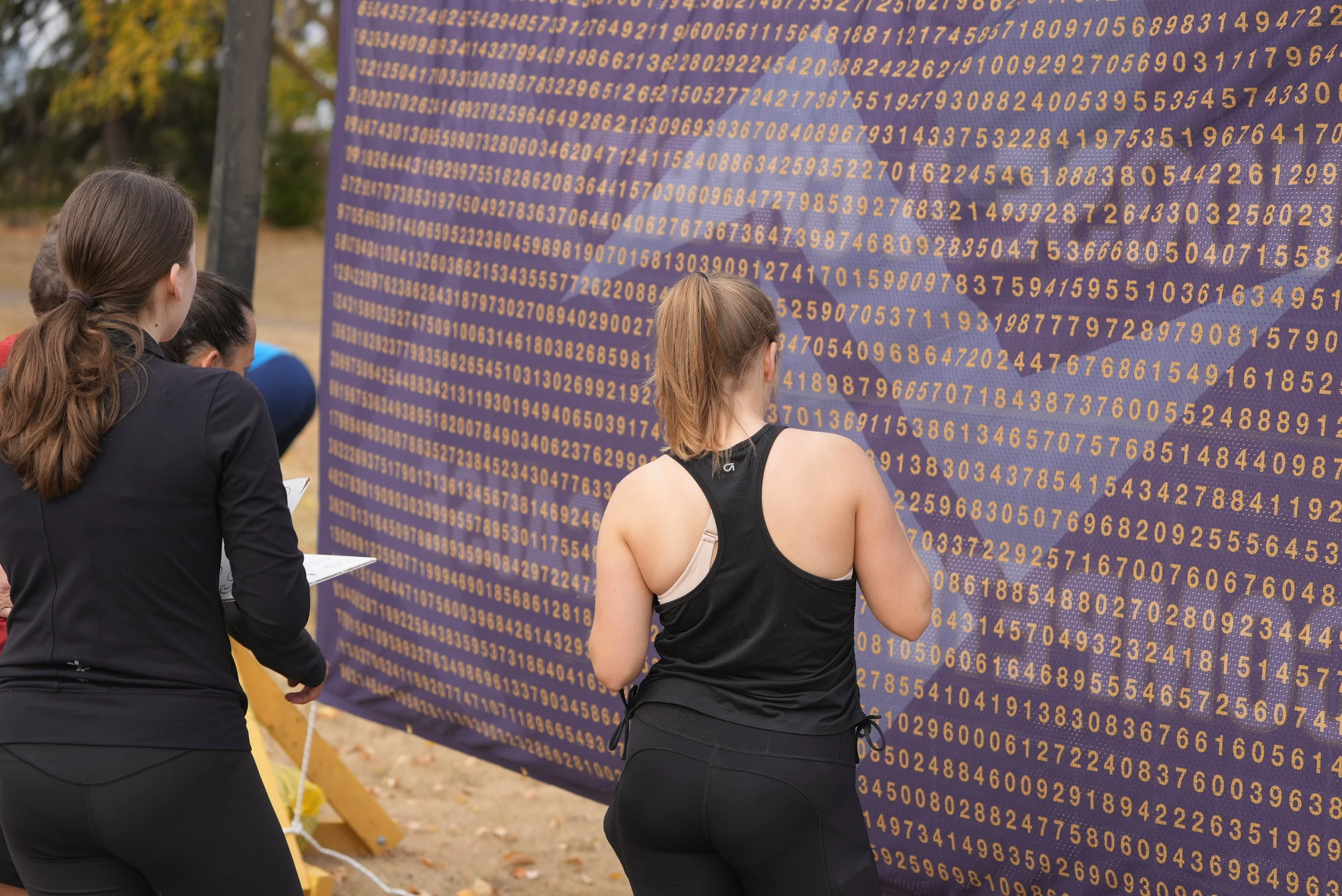 Three women stand outdoors in front of a large purple display covered with rows of yellow numbers, appearing to observe the display closely.