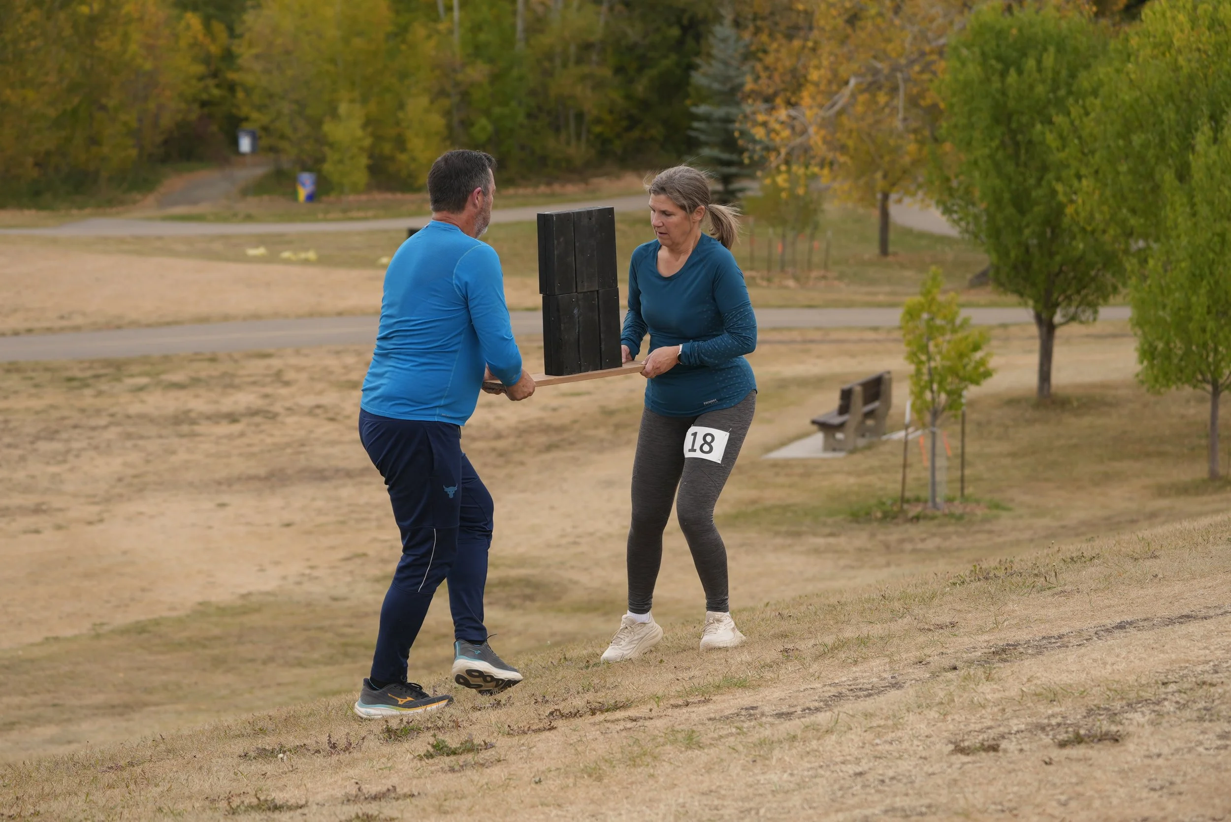A man and a woman exchange a wooden plank with bricks on it in a park with autumn trees. The woman has a race number 18 on her leggings.