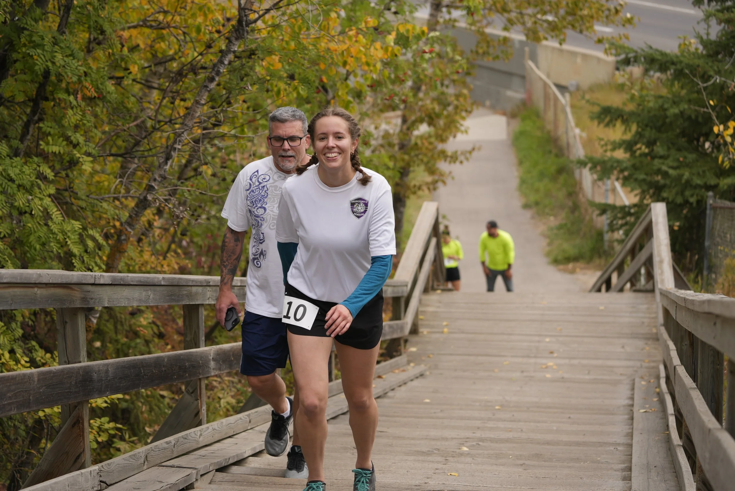 A young woman and an older man are running together on a wooden bridge in a park, smiling and enjoying a jog during autumn with trees showing fall foliage in the background.