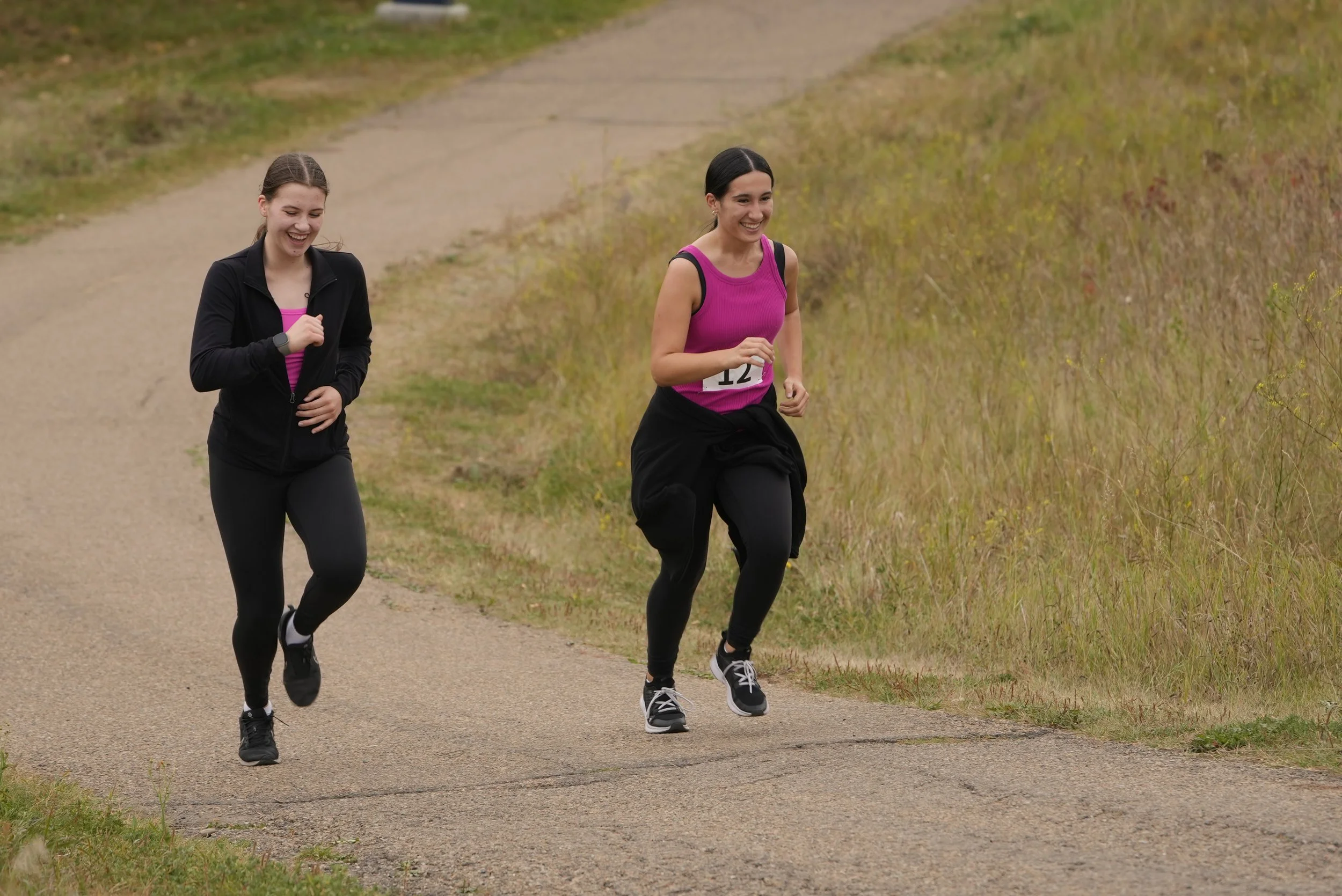 Two women running on a dirt trail in a natural outdoor setting, smiling and enjoying the activity.