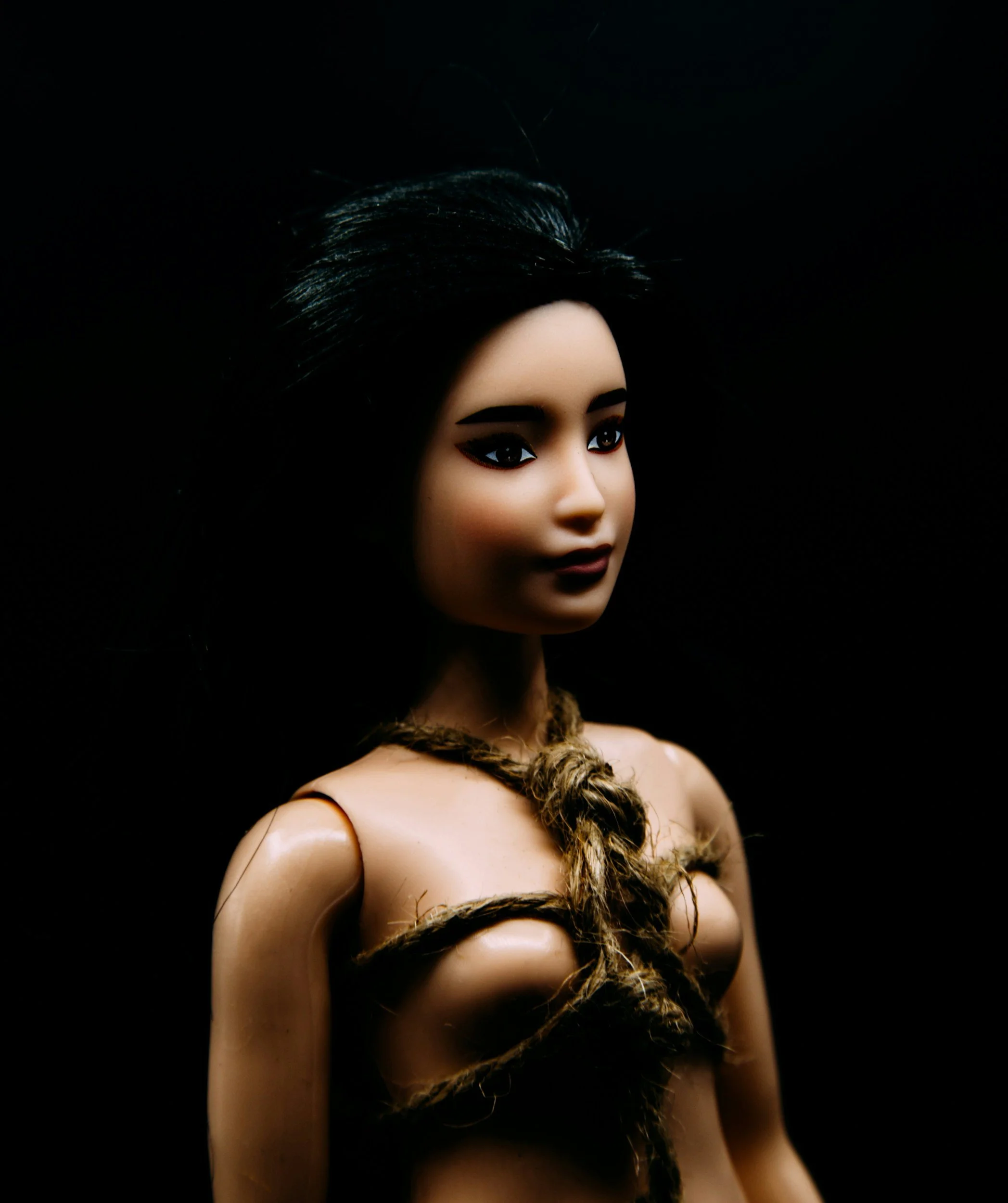 A female doll with dark hair, dark eyes, and dark lipstick, tied with rope around the shoulders and torso, with a plain black background.