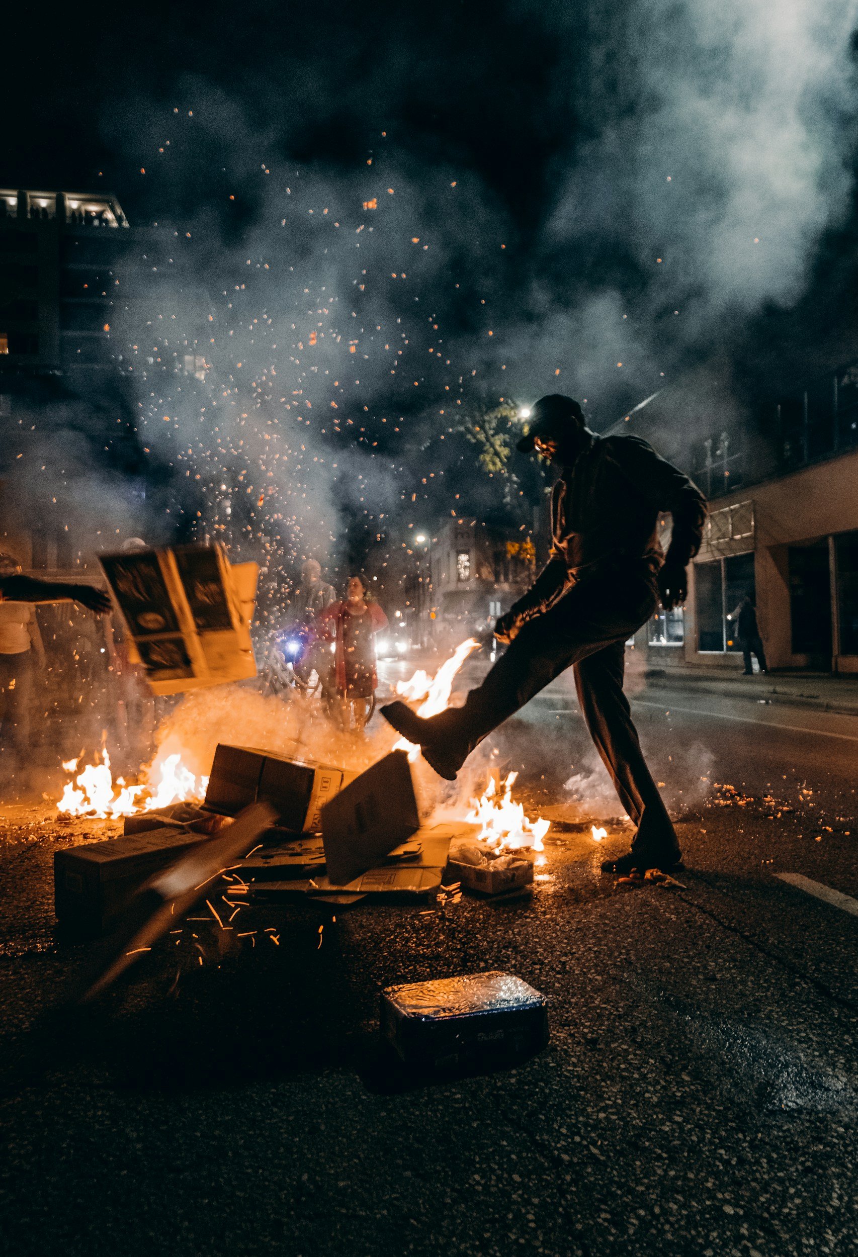 Person throwing a box into a fire on a city street at night, with smoke and sparks in the air and other people in the background.