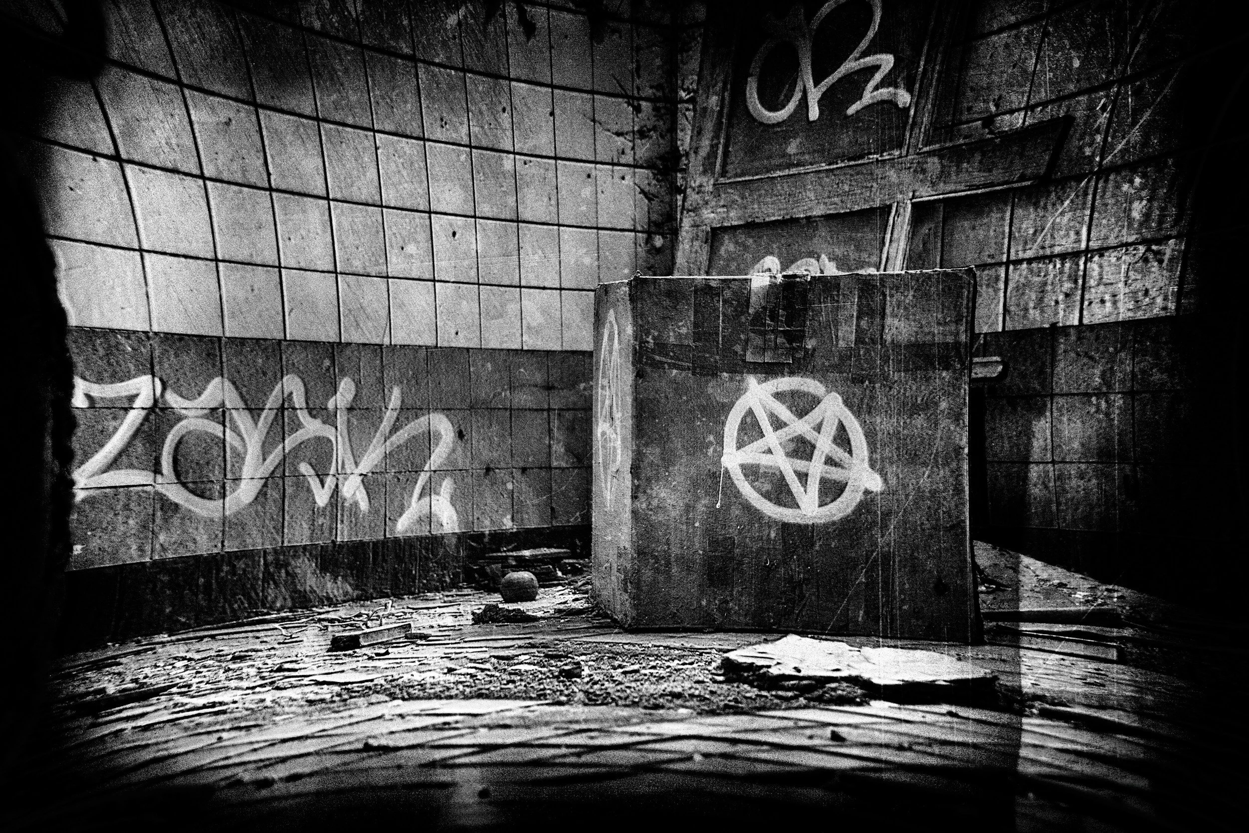 Black and white photo of an abandoned, dilapidated room with graffiti. A concrete block with a pentagram symbol spray-painted on it is in the foreground. The walls are covered with tiles, some of which are broken or missing, and more graffiti is visible on the walls.