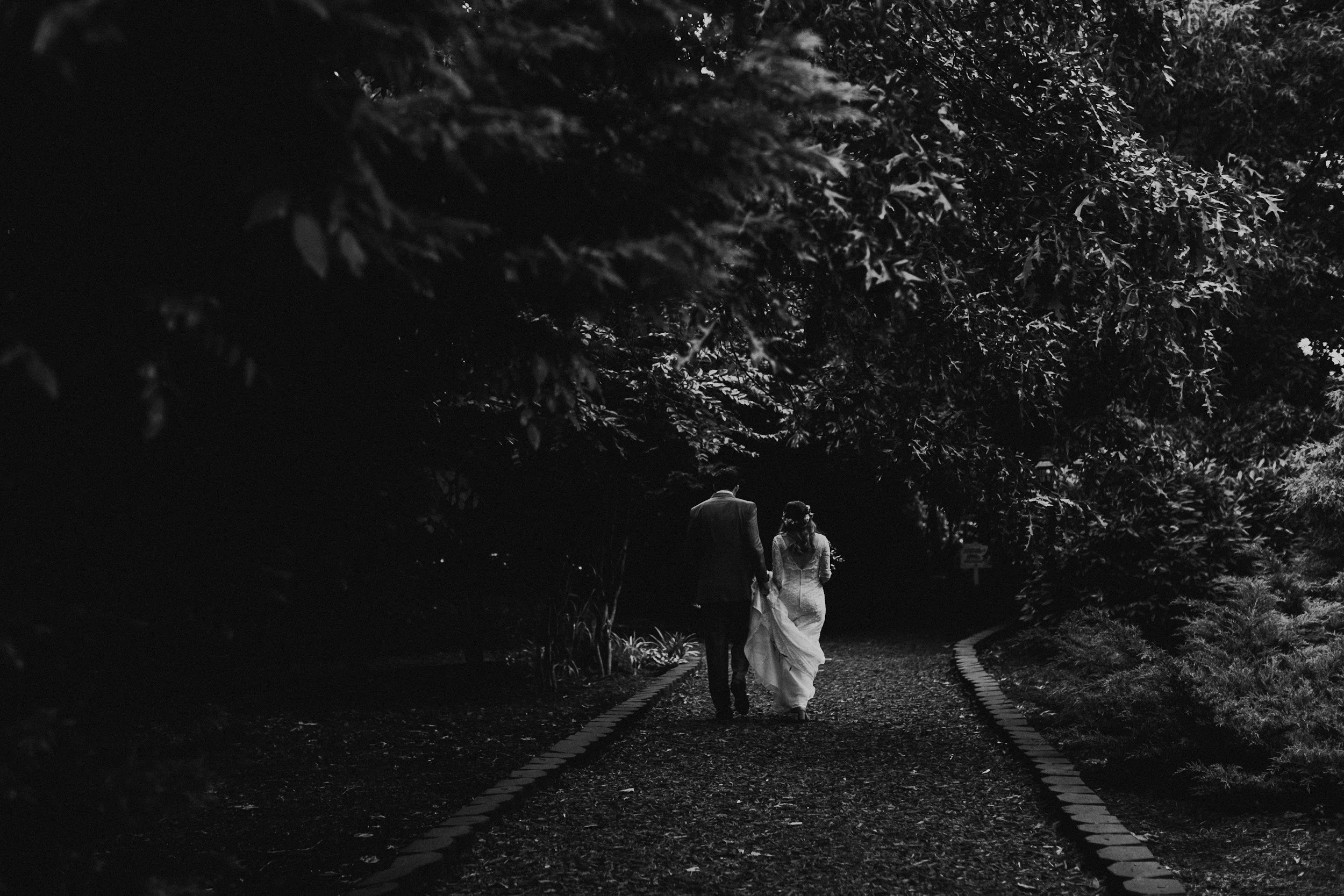 A black and white photo of a bride and groom walking hand in hand on a path surrounded by trees in a garden.