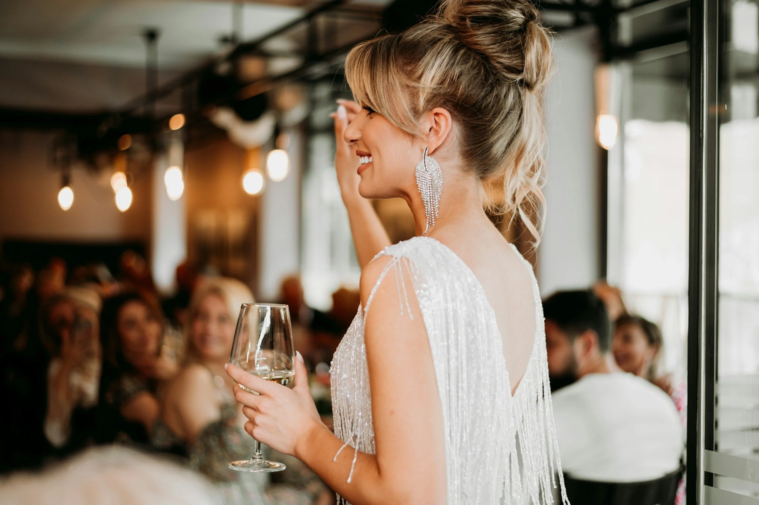 Bride wearing a beaded white dress and large chandelier earrings, smiling and holding a wine glass at her wedding reception.