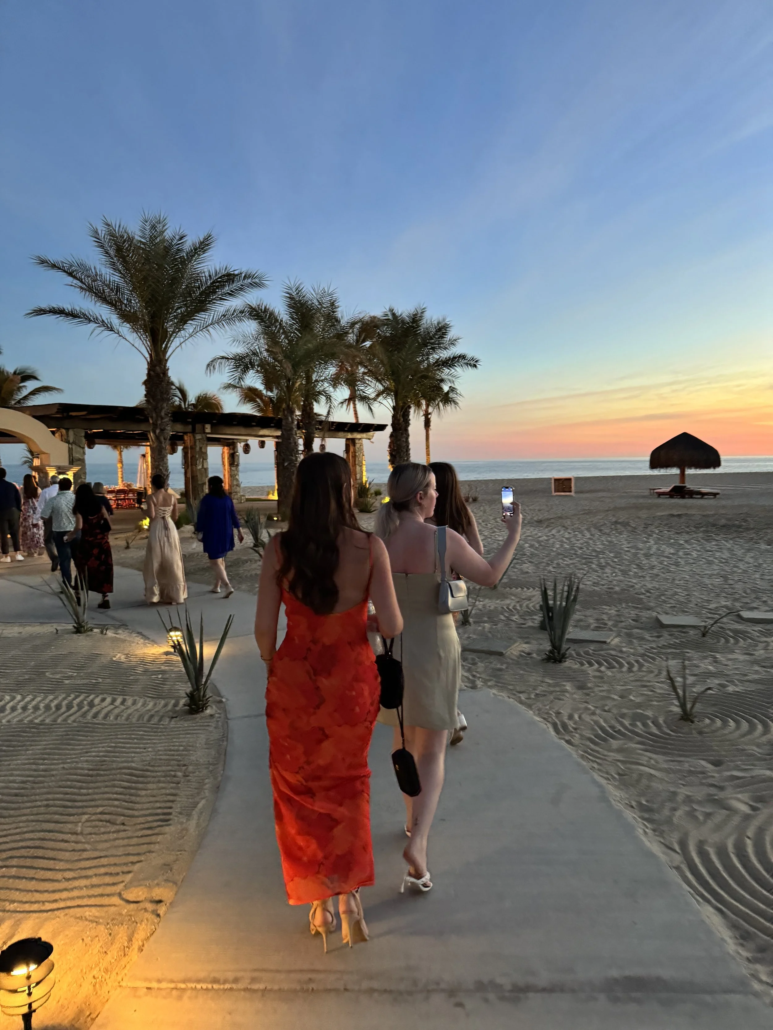 People dressed in summer attire walking on a beachside path during sunset, with palms, a thatched umbrella, and a beachside bar visible.