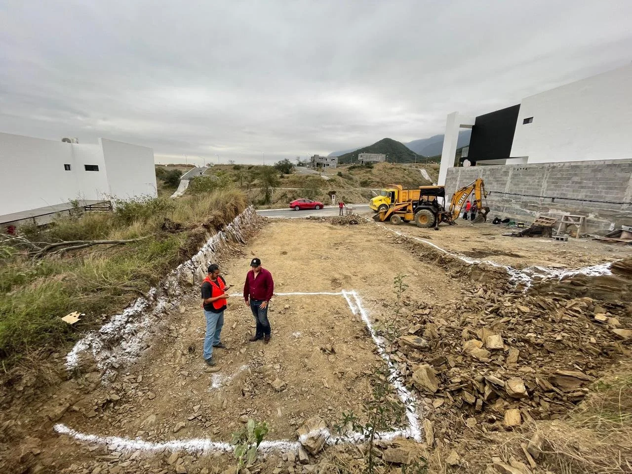 Sitio de construcción en un terreno excavado con marcas blancas en el suelo, maquinaria pesada y dos personas conversando.