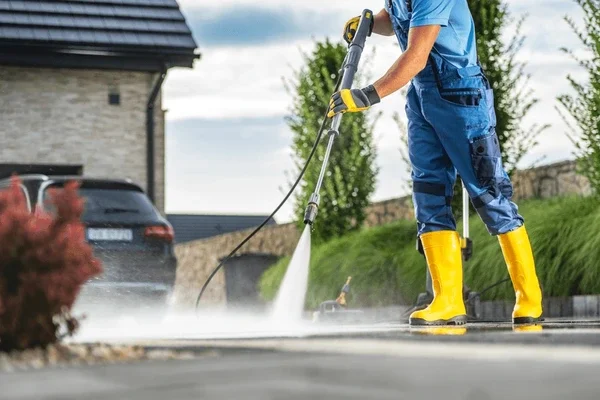 A worker in a blue uniform and gloves uses a robotic pool cleaner to clean a backyard swimming pool surrounded by a patio and landscaping.