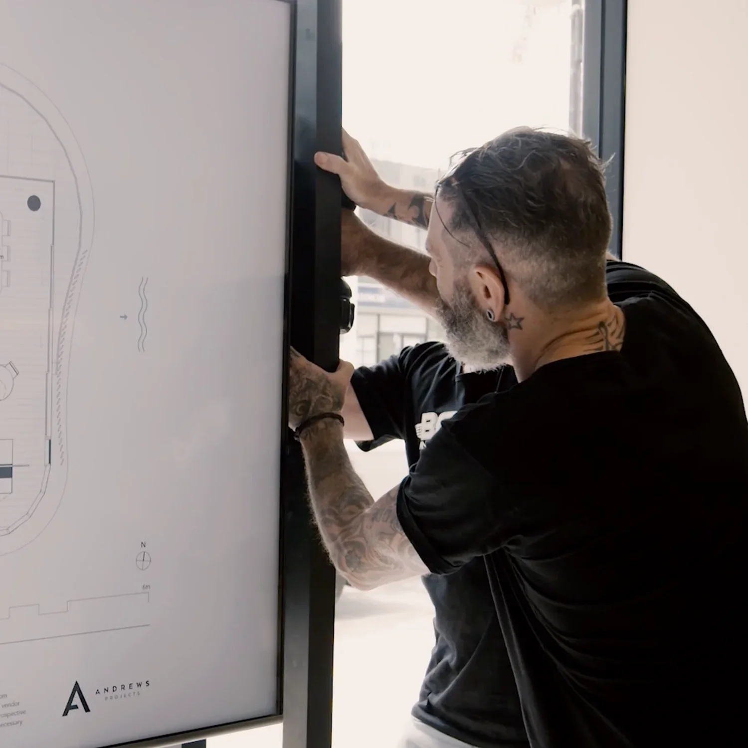 A man with tattoos and glasses, wearing a black shirt, is installing or adjusting a large digital screen in a bright room with windows behind him. Gold Coast