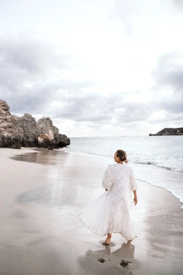 A woman in a long white dress walking barefoot along the sandy beach near rocky cliffs and ocean under a cloudy sky.