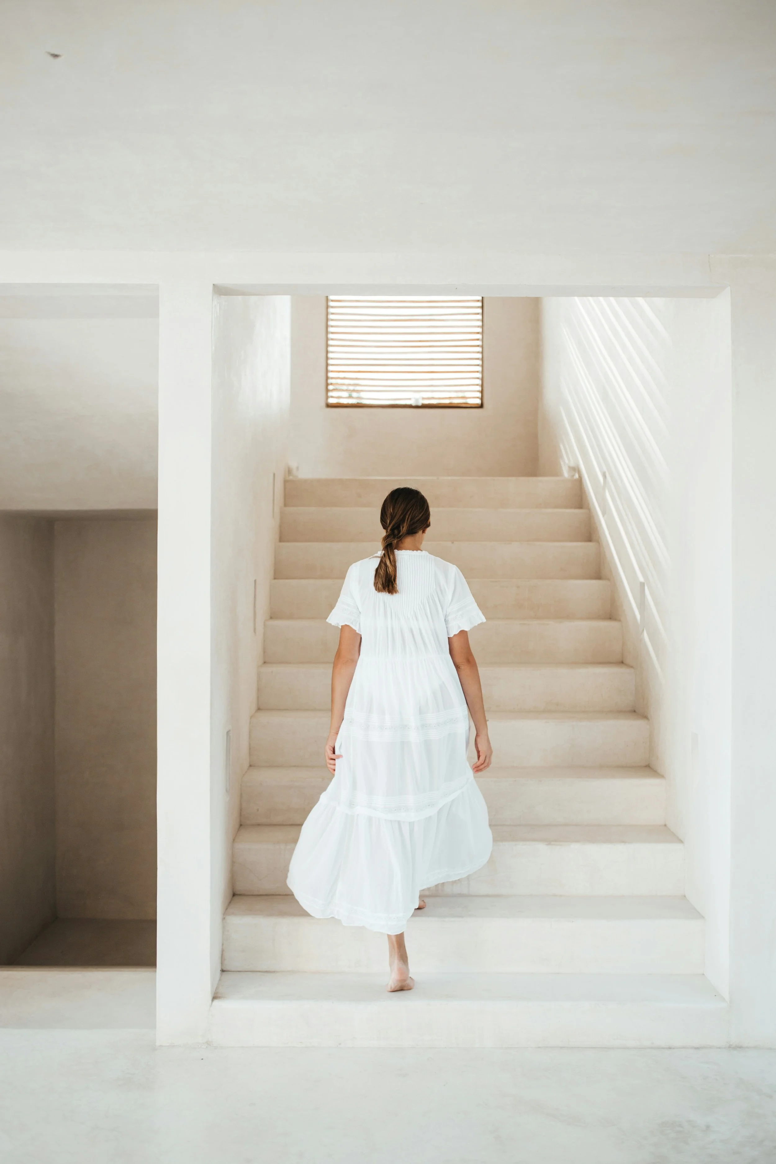 A woman in a white dress walking barefoot up a flight of stairs in a minimalist, white-walled interior with a window at the top letting in natural light.