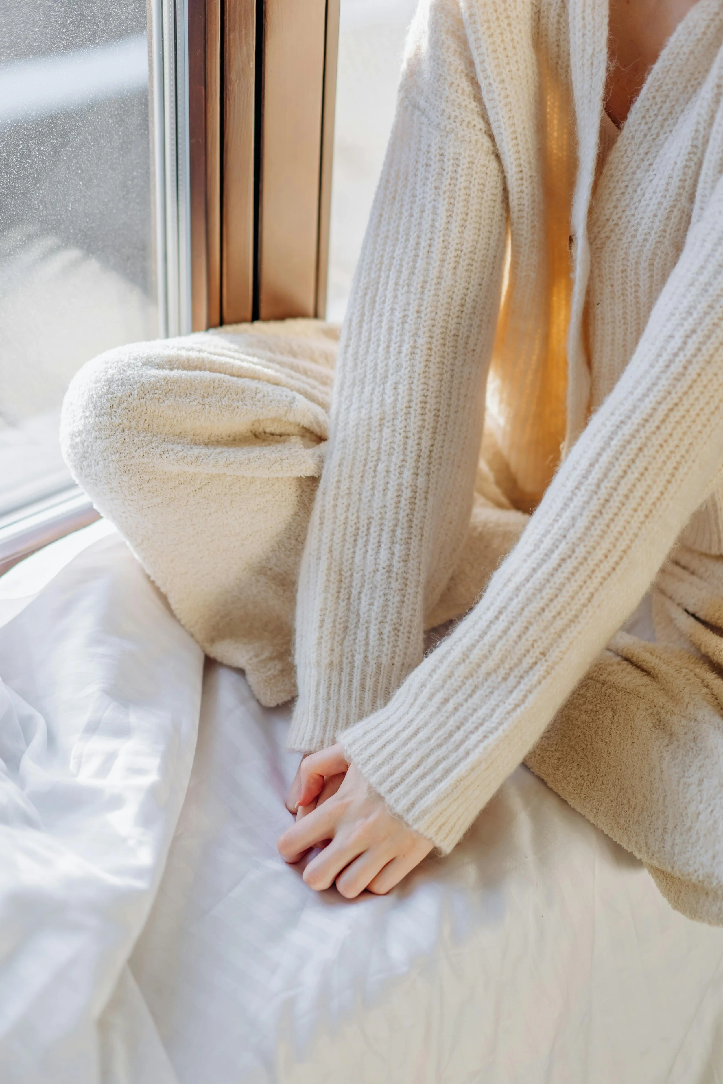 Person in a cream-colored, ribbed knit sweater sitting on a bed near a window with raindrops on the glass.
