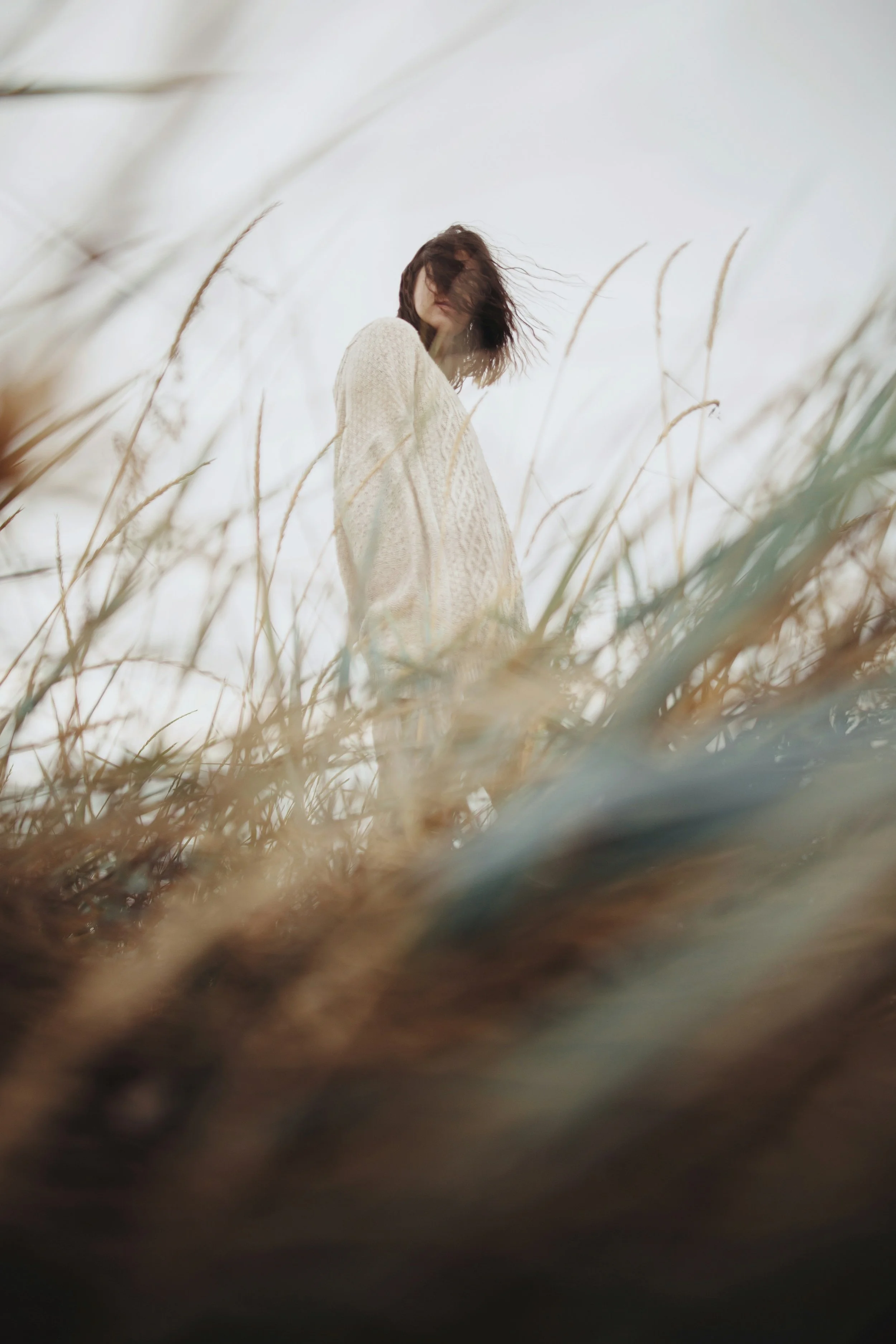 A woman standing in tall dry grass, wearing a cream-colored sweater, with her hair blowing in the wind against a cloudy sky.