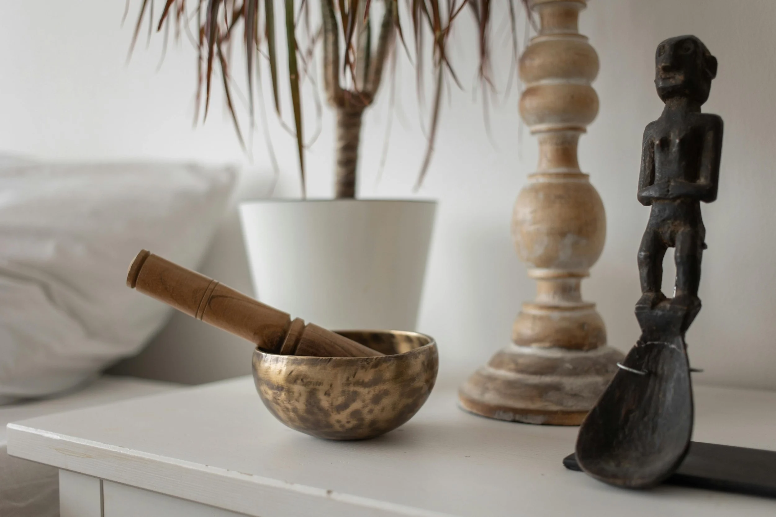 Decorative scene with a wooden pestle in a metallic bowl, a black carved figurine, a wooden candlestick, a potted plant with palm-like leaves, and a large white ceramic bowl, all placed on a white cabinet.