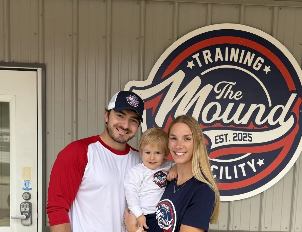 A smiling family of three stands in front of a large sign that reads "Training The Mound Established 2025 Facility." The father is wearing a red and white baseball shirt and a baseball cap. The mother has long blonde hair and is wearing a navy T-shirt with the same logo. The young boy is between them, smiling, and wearing a white shirt with the same logo.