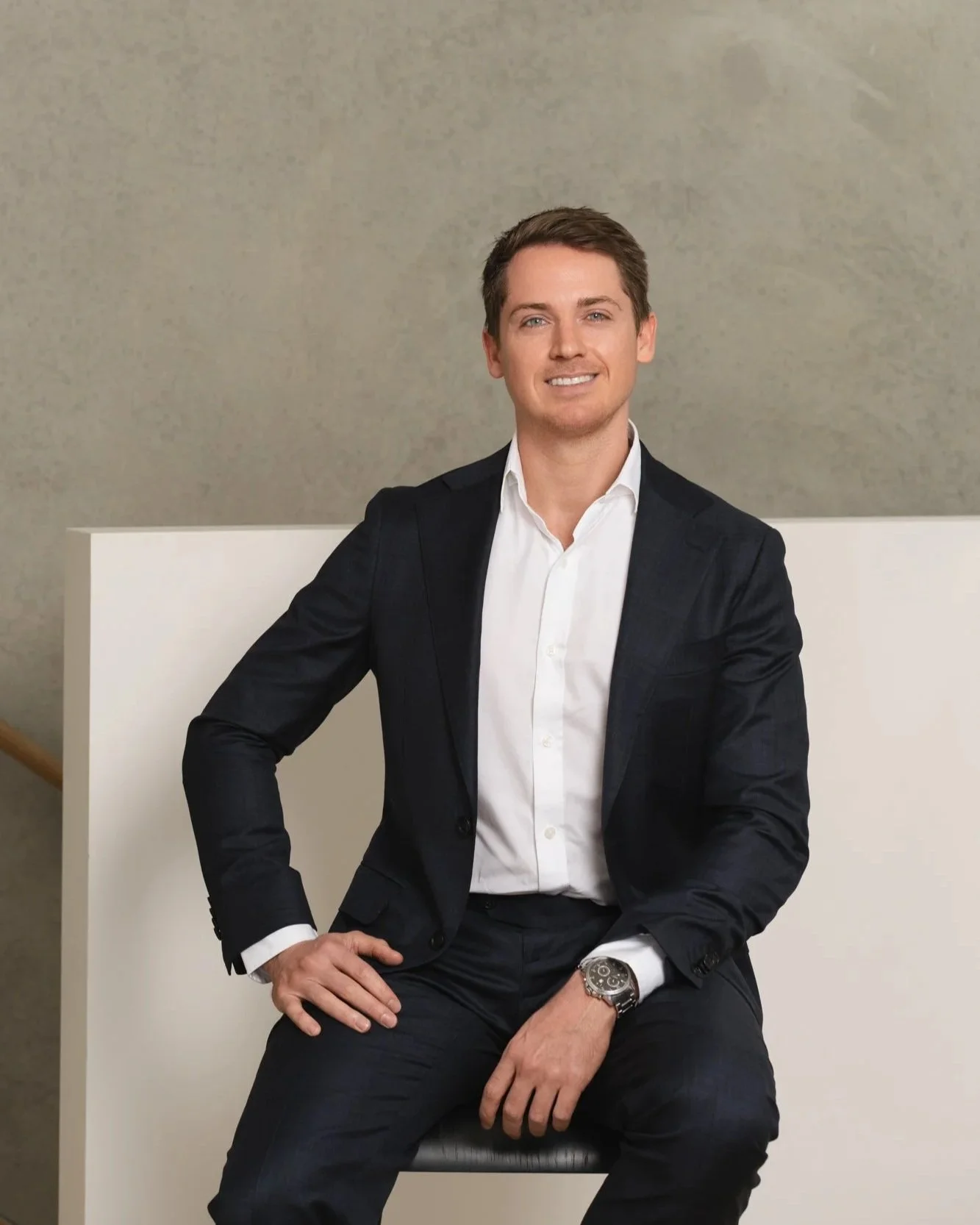 A man in a suit sitting on a chair against a neutral background, smiling at the camera.