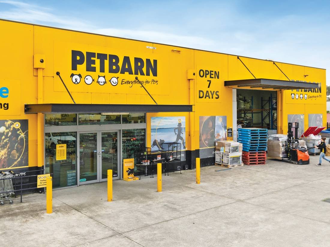 Exterior of a bright yellow Petbarn store with signage displaying the store's name and logo, front entrance with glass doors, and outdoor shopping carts. There are yellow bollards in front and a forklift to the side, with various stacked pallets and