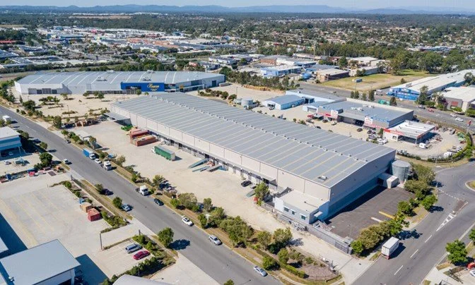 Aerial view of a large industrial warehouse with parking lot, trucks, and surrounding commercial area in an urban setting.