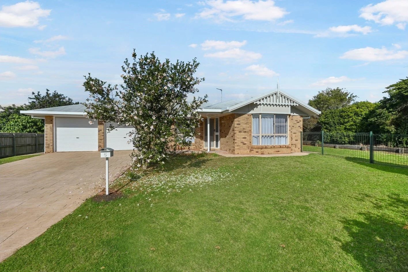 Front view of a single-story brick house with a white garage door, a small front yard with grass and a tree, a driveway, and a fence on the right side.