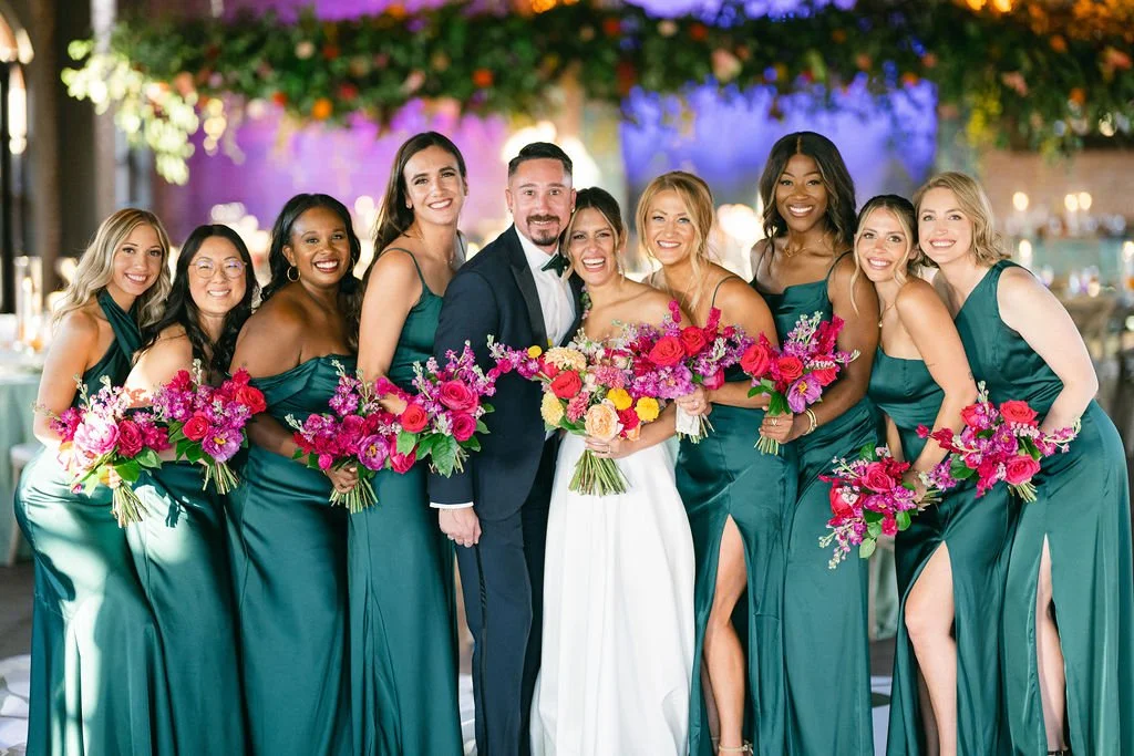 Group of women and a man at a wedding, with the women dressed in teal bridesmaid dresses and the man in a tuxedo, all holding bouquets of colorful flowers, in a decorated venue with hanging flowers and lights.