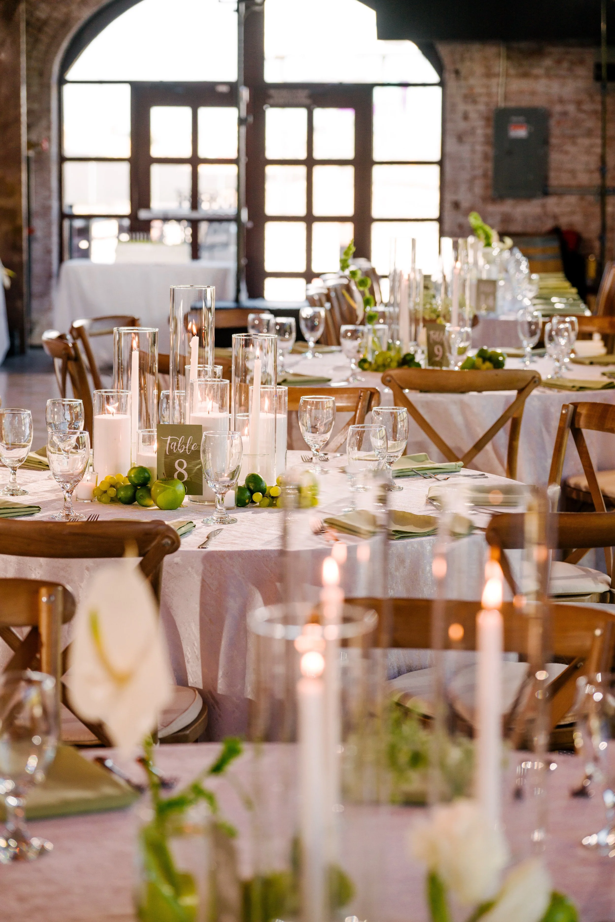 A decorated banquet table inside a rustic venue with exposed brick walls and large windows, featuring candles, glassware, and green accents for an event.