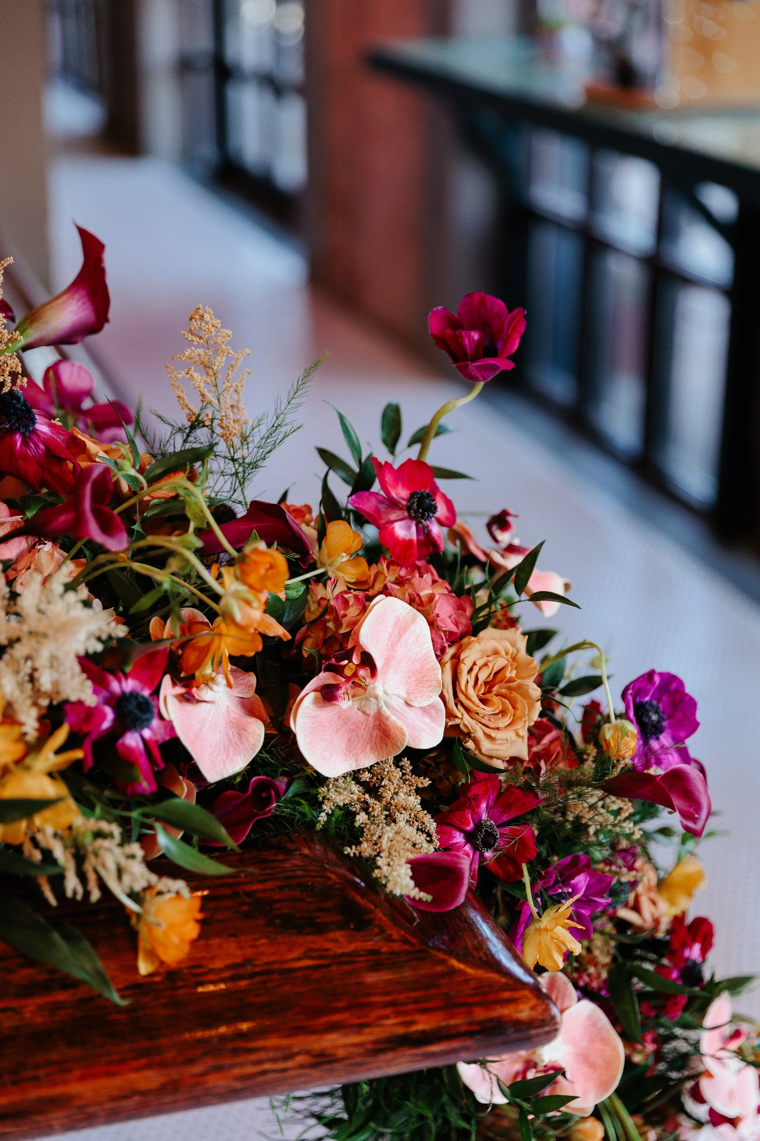 A colorful flower arrangement with pink, orange, purple, and cream flowers, placed on a wooden surface near a window with a black railing in the background.