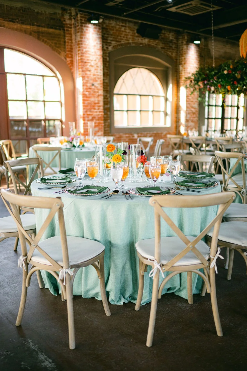 A round banquet table decorated with a light green tablecloth, set with green plates, silverware, glasses of water and juice, and small floral arrangements with yellow, orange, and red flowers, located in a rustic indoor venue with exposed brick walls and large arched windows.