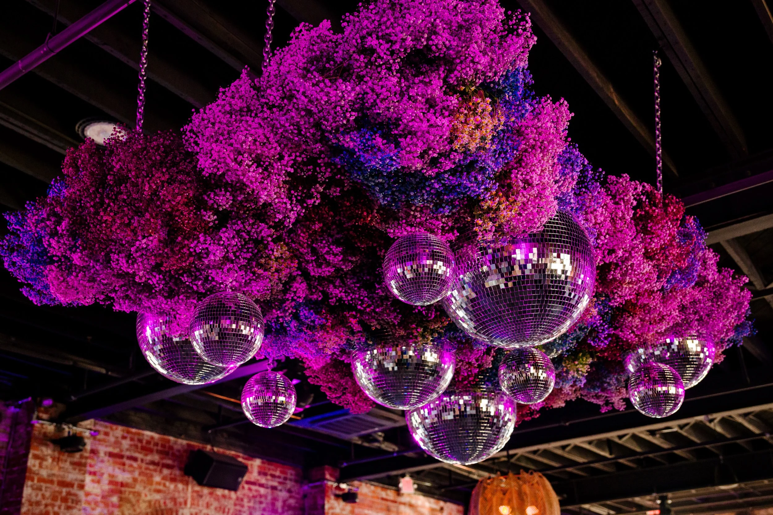Hanging pink and purple floral arrangement with multiple disco ball ornaments in a dark indoor space with brick walls and a high ceiling.