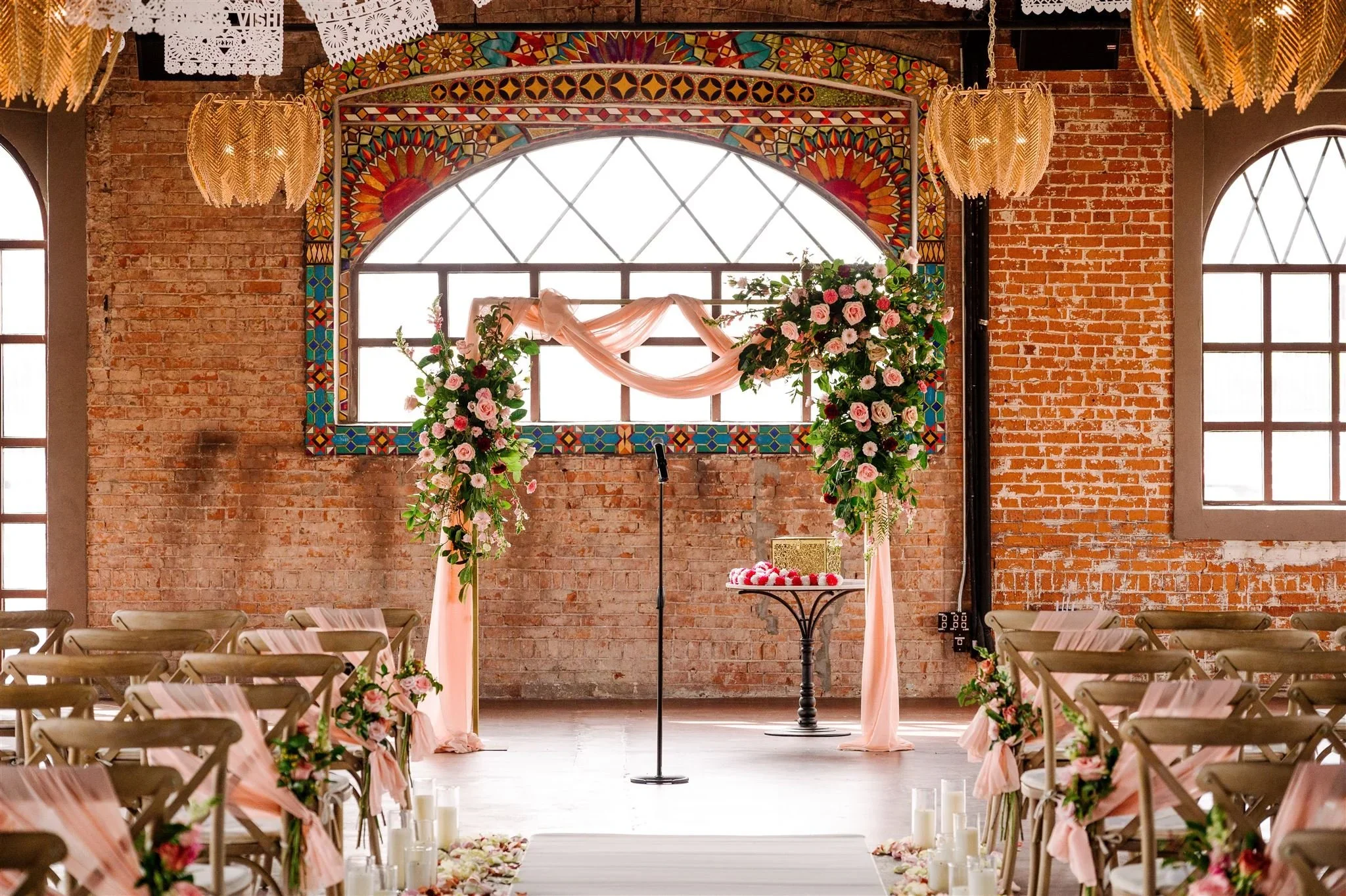A wedding setup with rows of chairs decorated with pink ribbons leading to an altar with flowers and pink fabric draping, in a venue with brick walls and large windows.