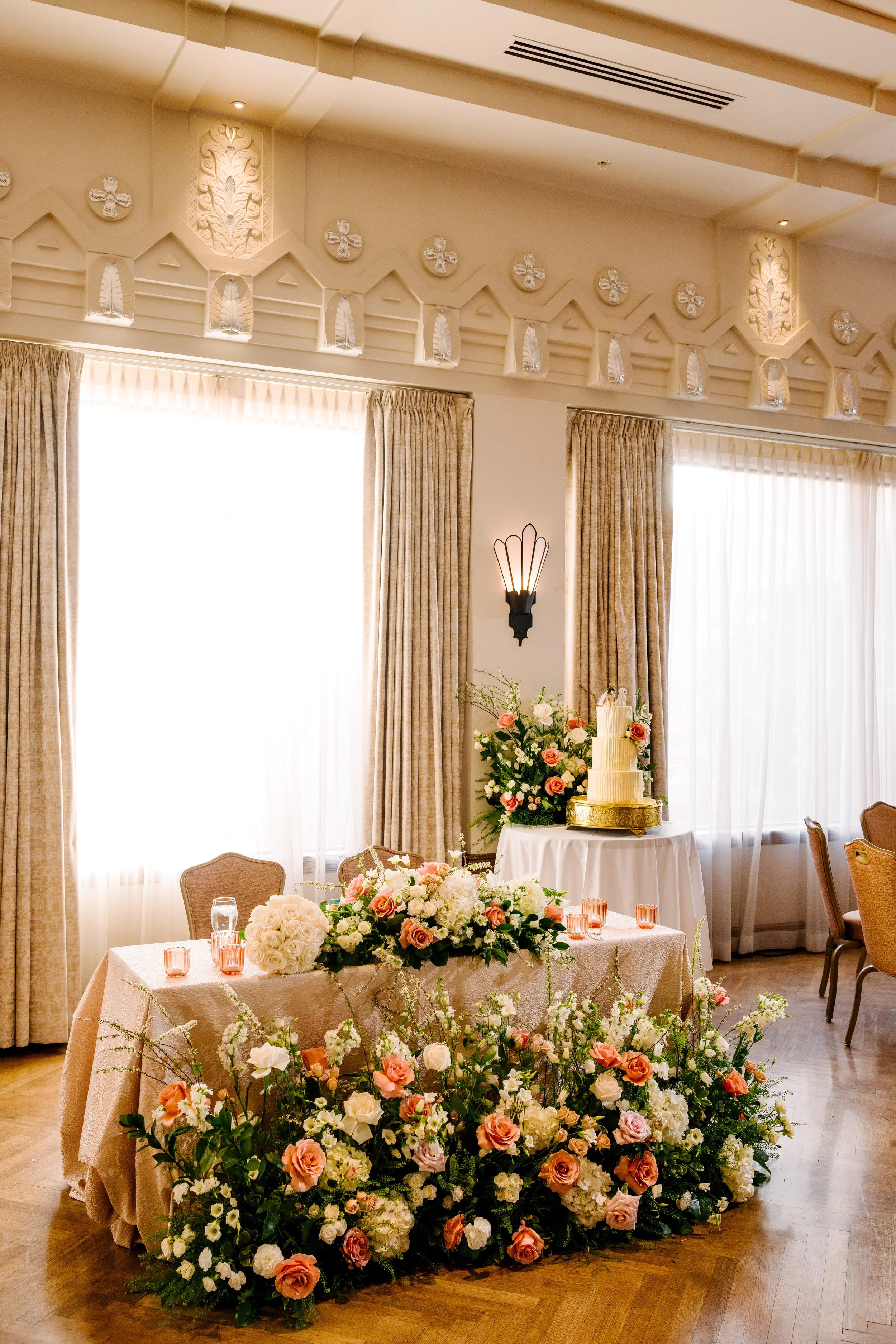 Elegant banquet room decorated for a wedding or celebration with a floral centerpieces, a cake, and topiary decor on the walls.
