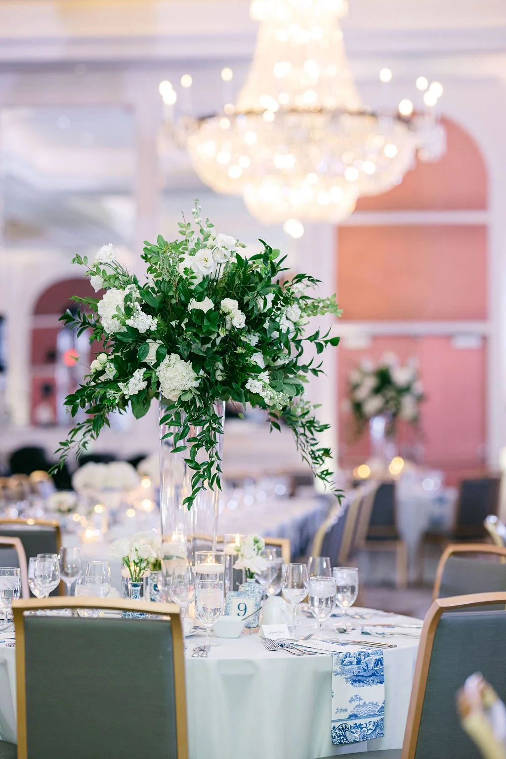 Elegant event venue with table featuring a tall floral centerpiece of white flowers and green leaves, set with glassware and tableware, chandelier overhead, and pink paneled walls in the background.