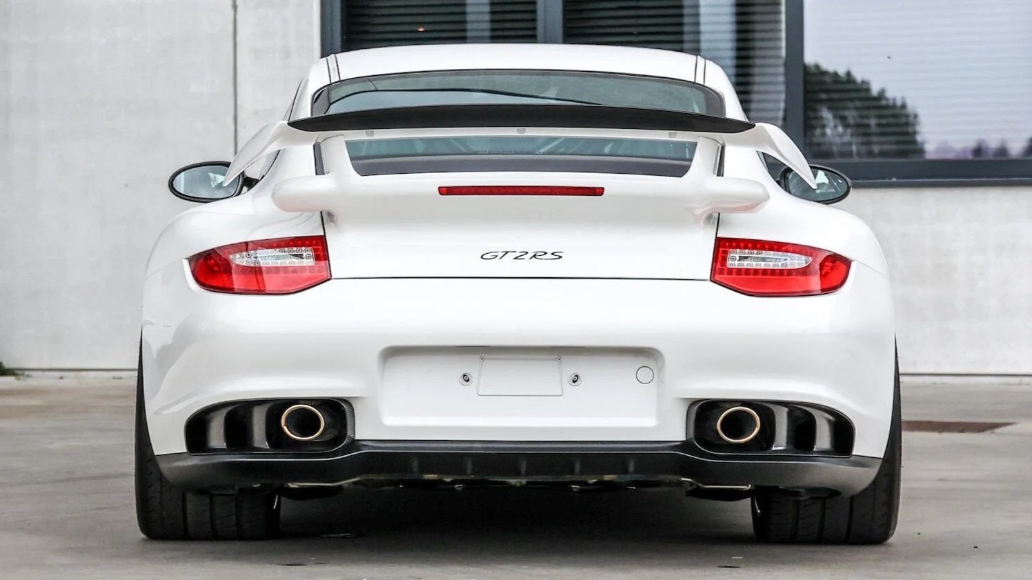 Rear view of a white Porsche 911 GT2 RS sports car parked on concrete with a modern building with glass windows in the background.