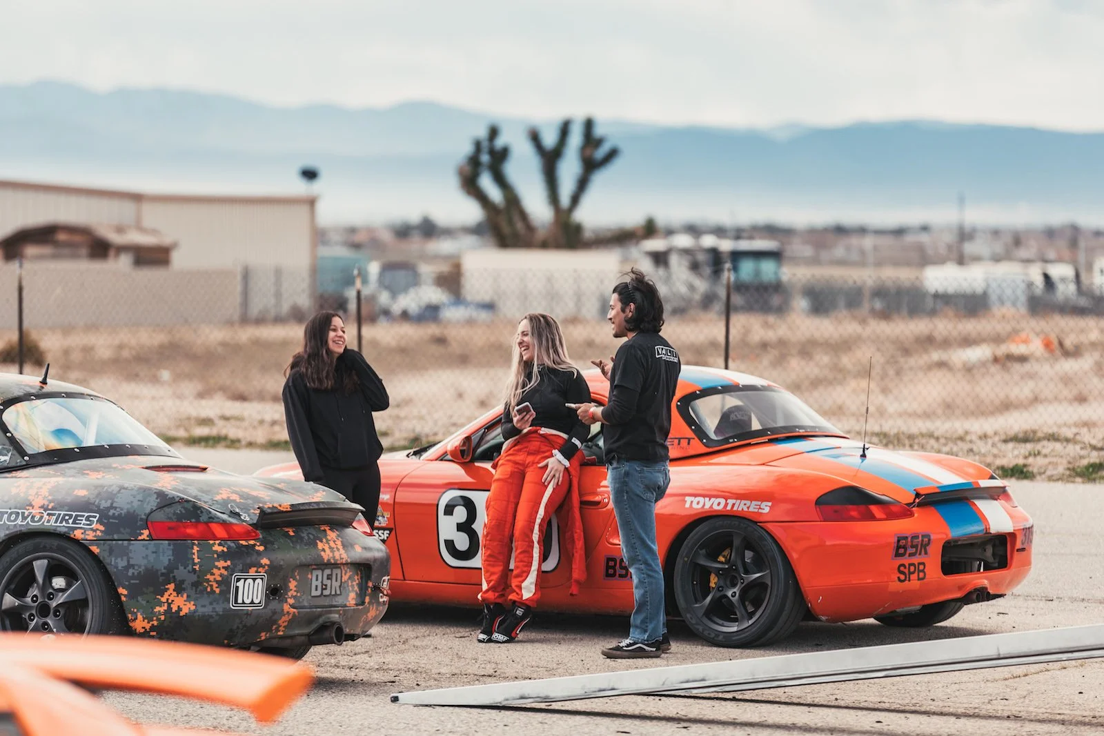 Three people standing next to race cars in a desert area, smiling and talking.